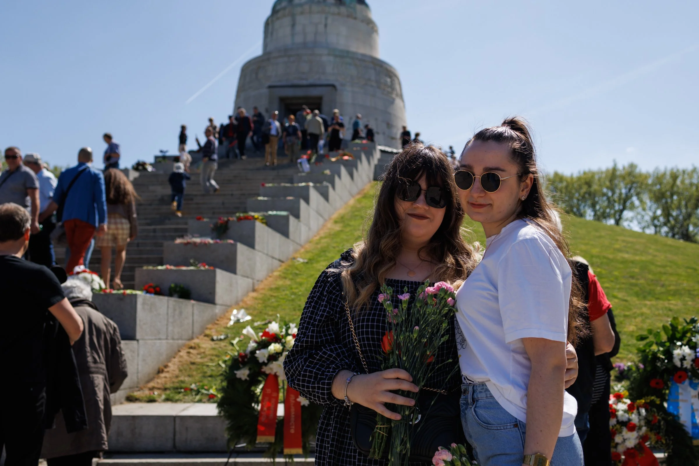 Two women with sunglasses holding flowers, standing in front of a memorial with many people and floral arrangements around it, under a clear blue sky.