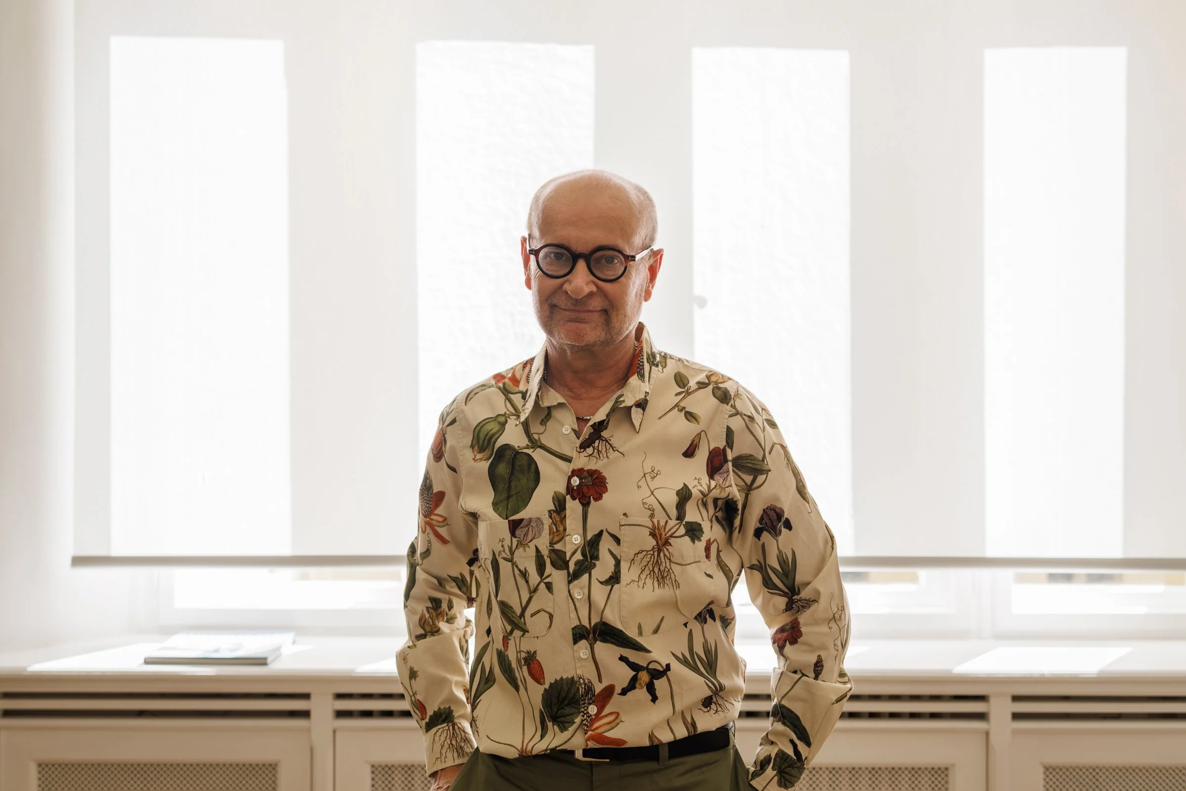 A man with glasses and a bald head wearing a beige shirt with floral and botanical prints stands in front of a window with vertical blinds, with books on a windowsill behind him.
