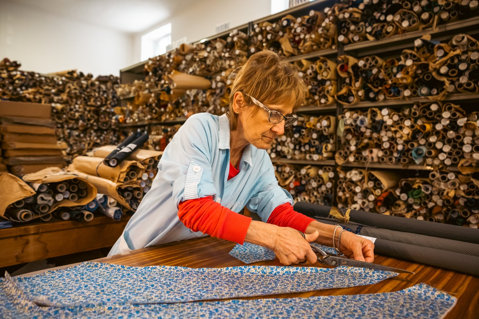 Female artisan meticulously cutting patterned blue fabric with shears at a workbench in the Petronius workshop.