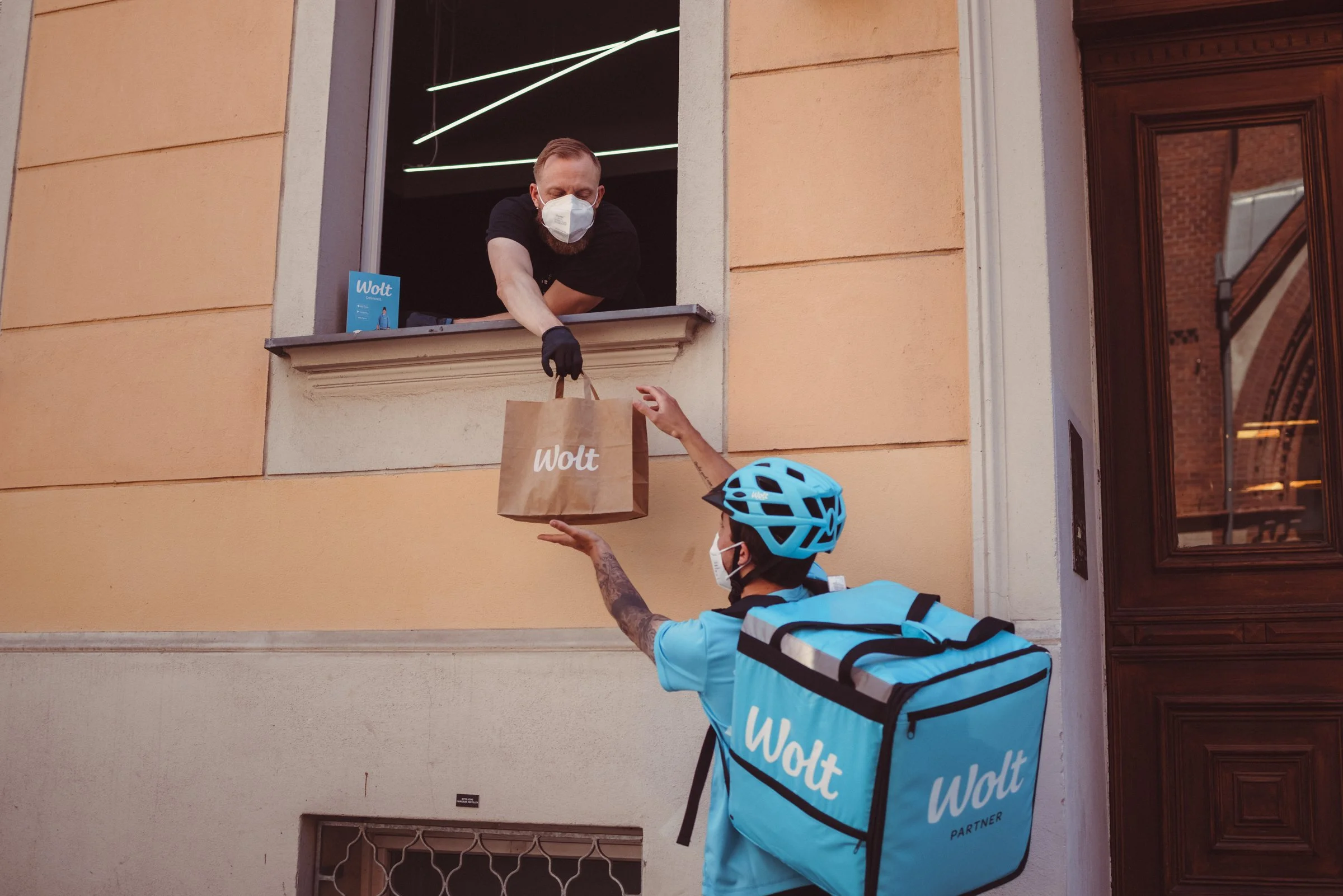 A person on a bicycle, wearing a blue helmet and blue Wolt delivery uniform, reaches up to receive a package from a man at a window. The delivery man, wearing a face mask and black gloves, is handing down a paper bag from the window of a peach-colore