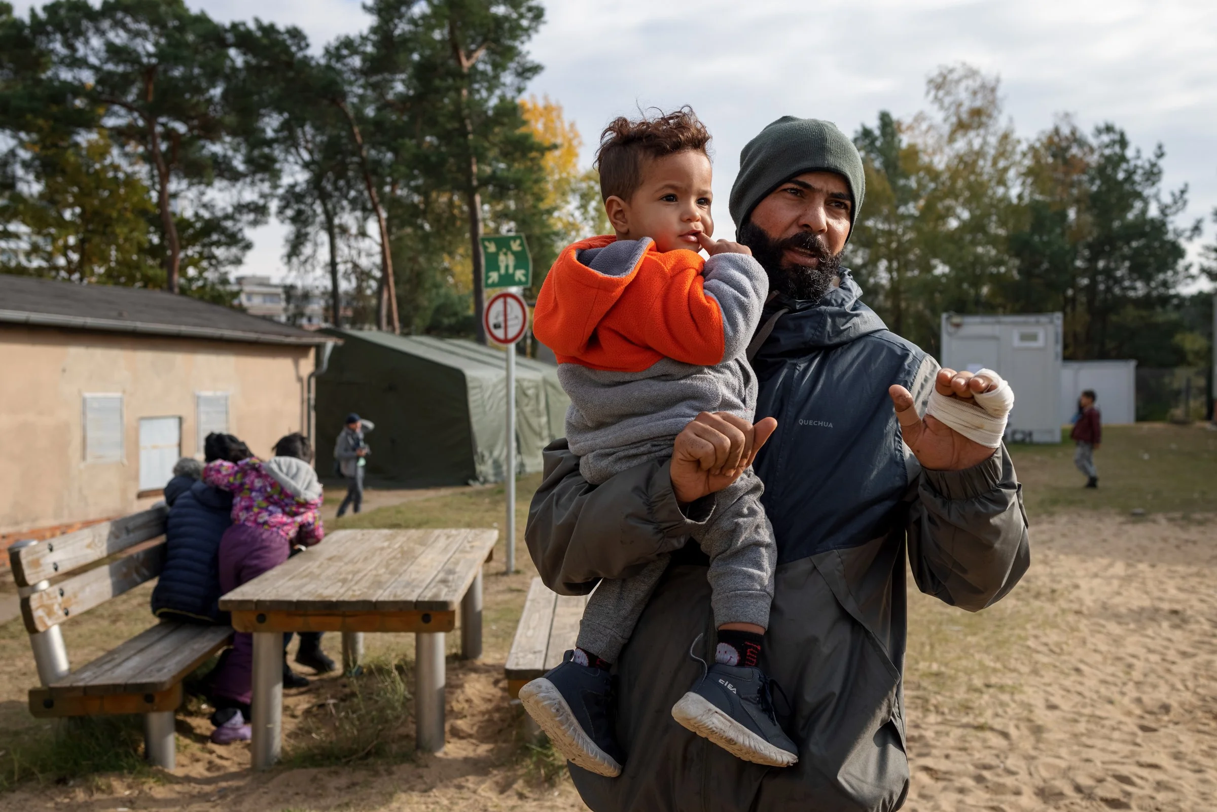 A man with a beard, wearing a gray beanie and a dark jacket, is carrying a young boy with curly hair, dressed in an orange and gray jacket, on his back. They are outdoors in a park-like setting with trees, benches, and several other people in the bac