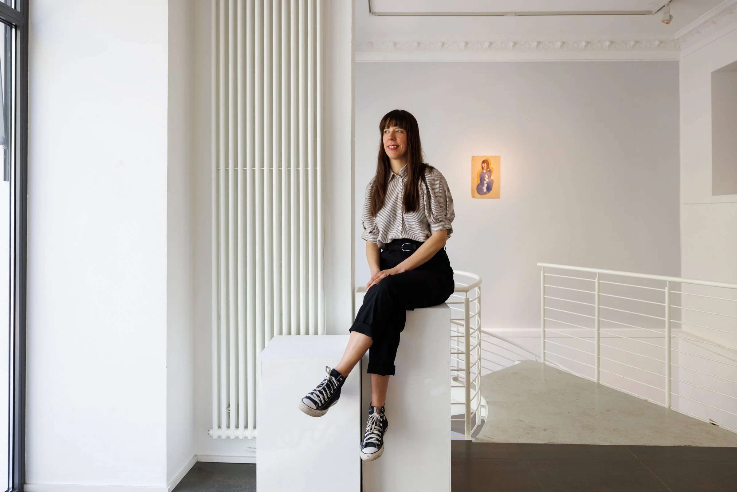A woman with long brown hair wearing a striped blouse, black pants, and black sneakers, sitting on a white architectural installation in a minimalist, modern gallery space. The gallery has white walls, a painting on the right wall, and a spiral stair