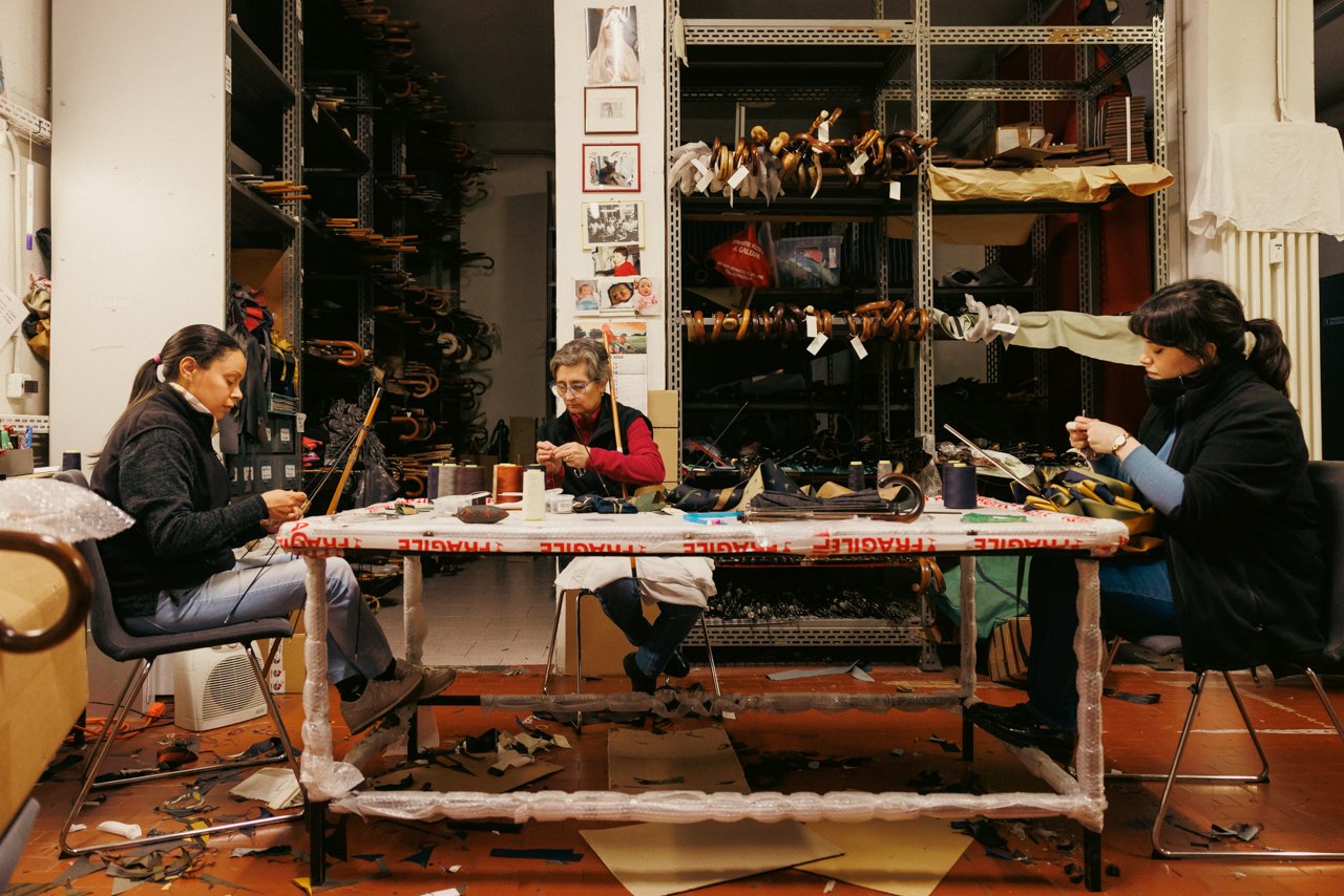Wider shot of the Maglia workshop floor with artisans working at tables amidst materials and tools.