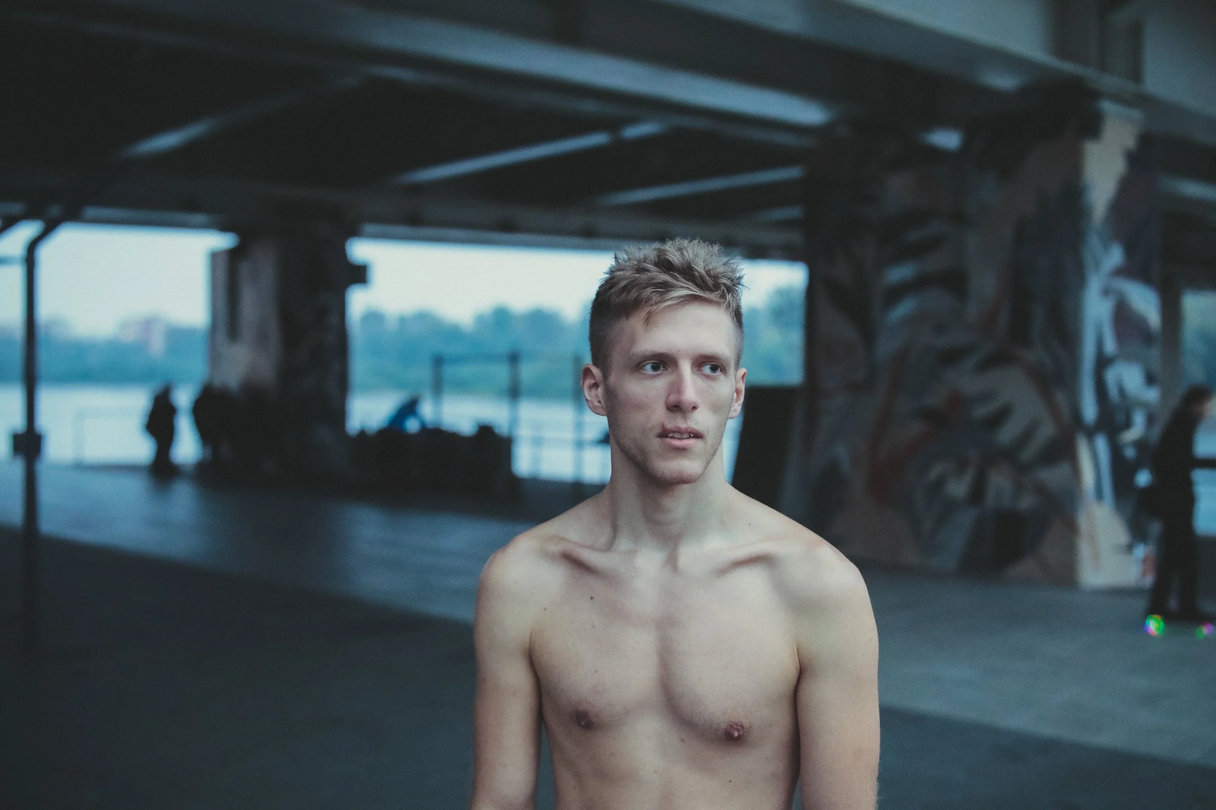 A shirtless young man, Linus Lewandowski, with short light brown hair stands under a bridge or overpass, with a background of water and a few people blurred in the distance, during dusk.