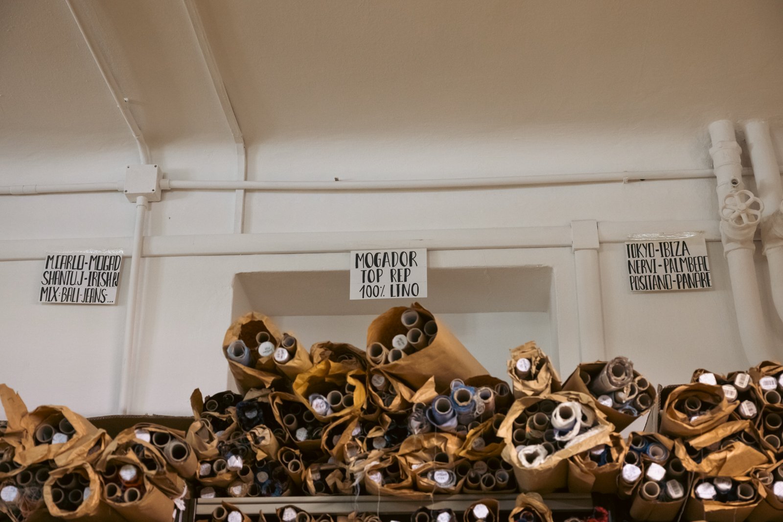 Storage shelf holding numerous rolls of labelled fabrics, including Mogador, Poplin, and Linen, at the Petronius atelier.