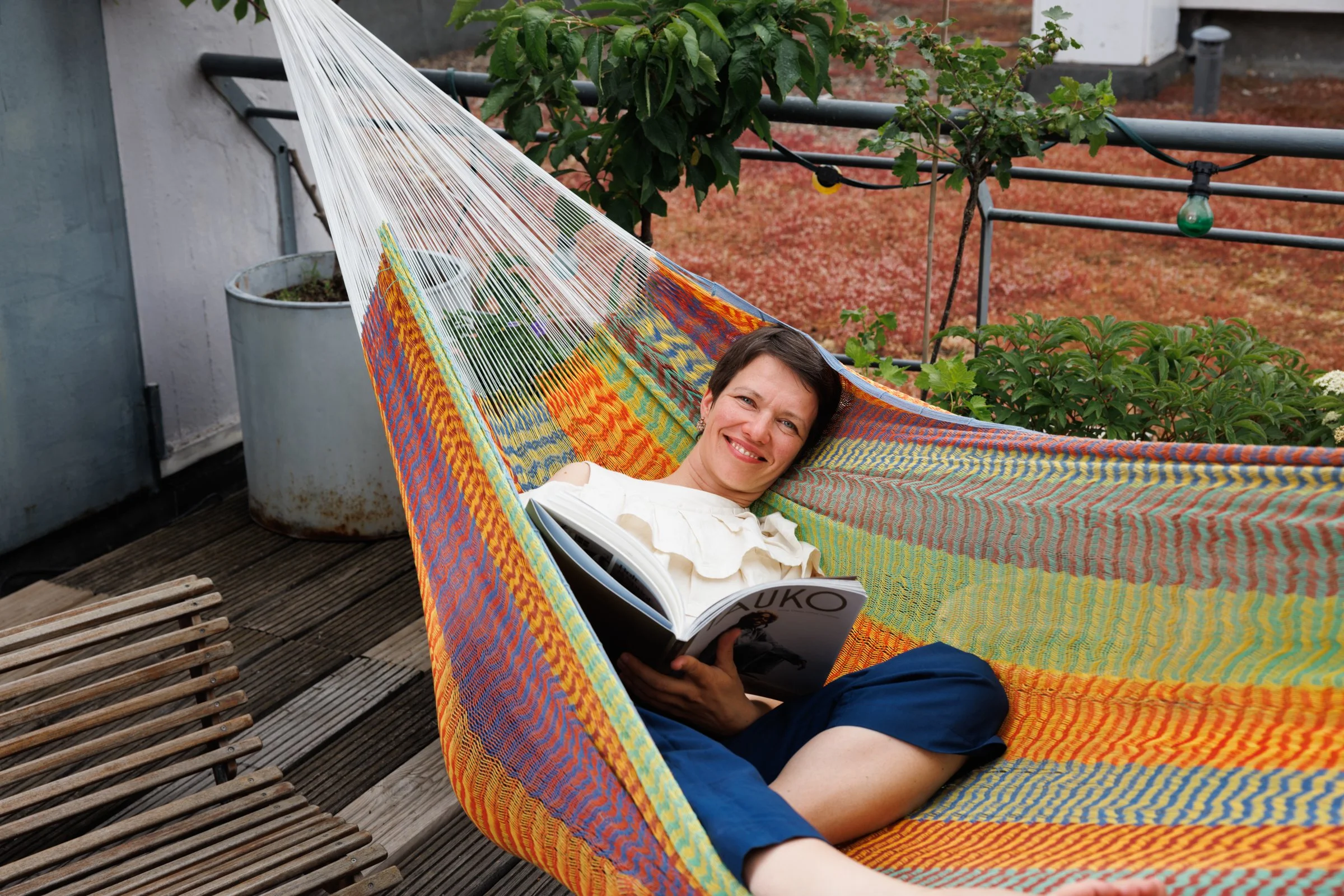 A woman is lying in a hammock on a rooftop garden, smiling and reading a magazine, with plants and string lights visible in the background.