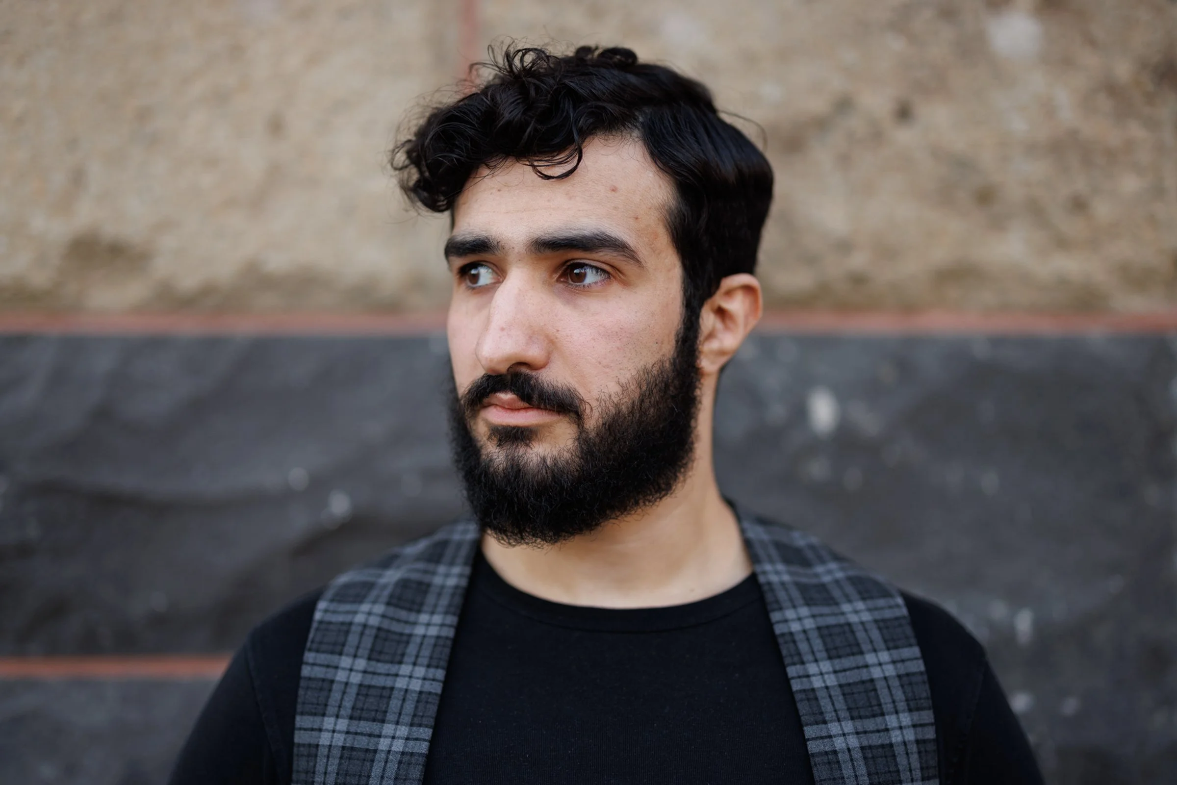 A young man with dark curly hair and a beard, wearing a black shirt and a checkered jacket, standing outdoors against a blurred background.