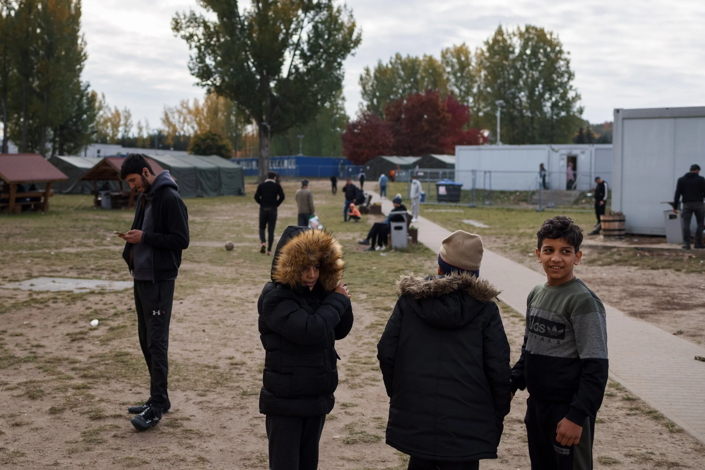 People gathered outdoors in a park-like setting with tents and trees, some standing, sitting, and talking.