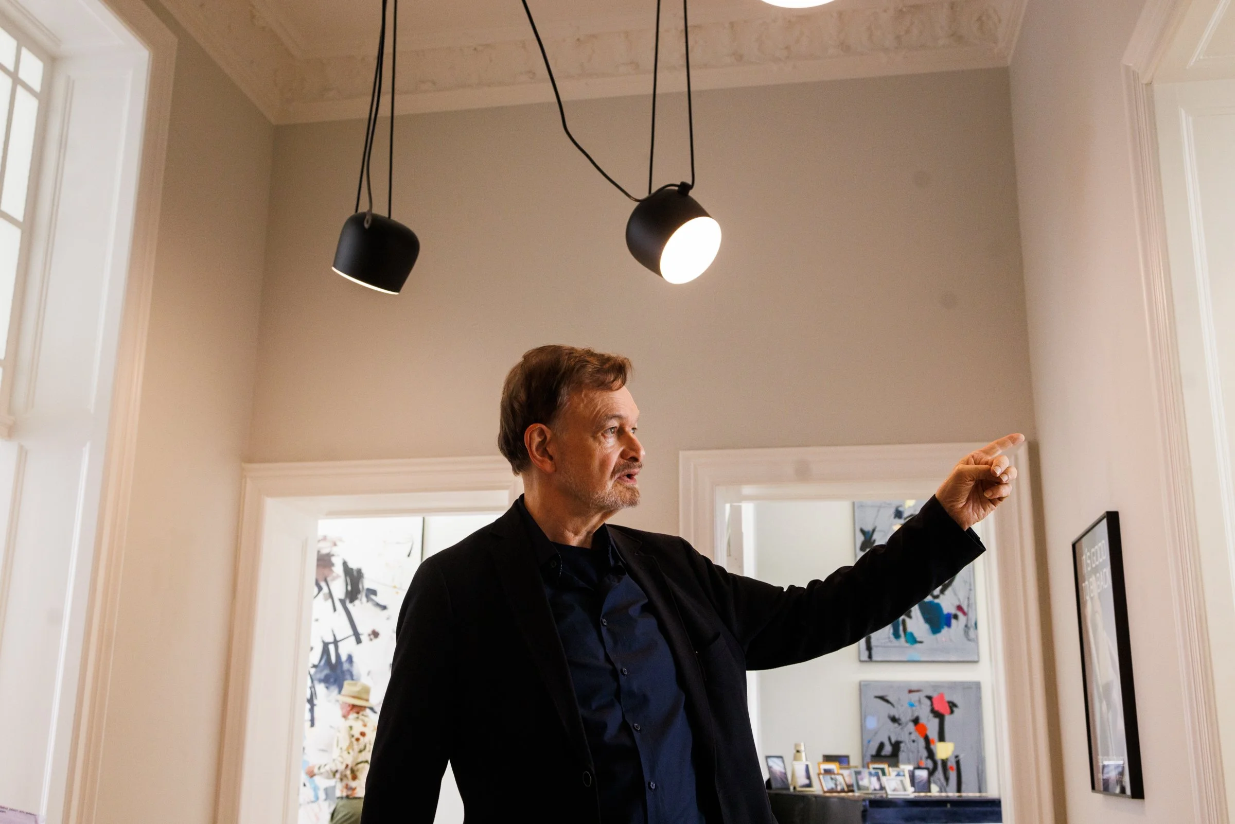 A man in a black jacket pointing and speaking indoors, with artwork and picture frames in the background.