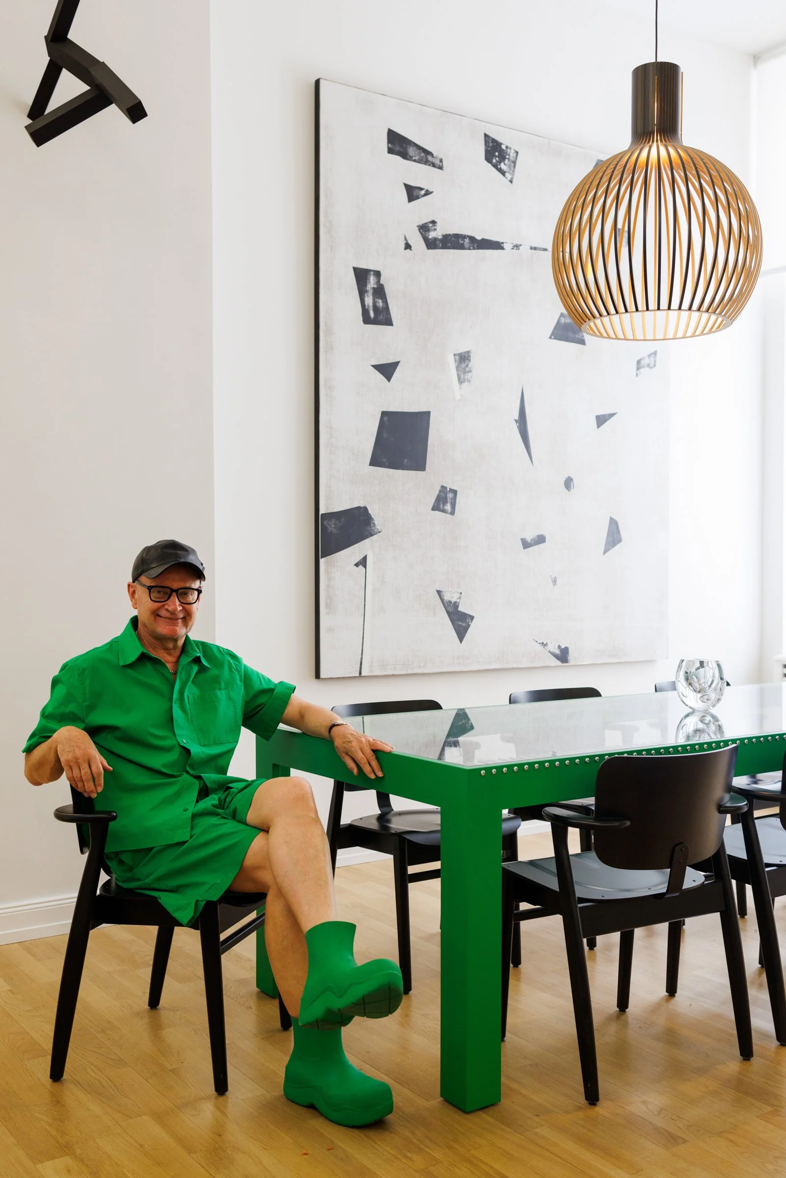 A man dressed in bright green clothing, including shorts, shirt, socks, and shoes, sitting at a modern green dining table in a room with minimalist decor. He is wearing glasses and a grey cap, smiling at the camera.