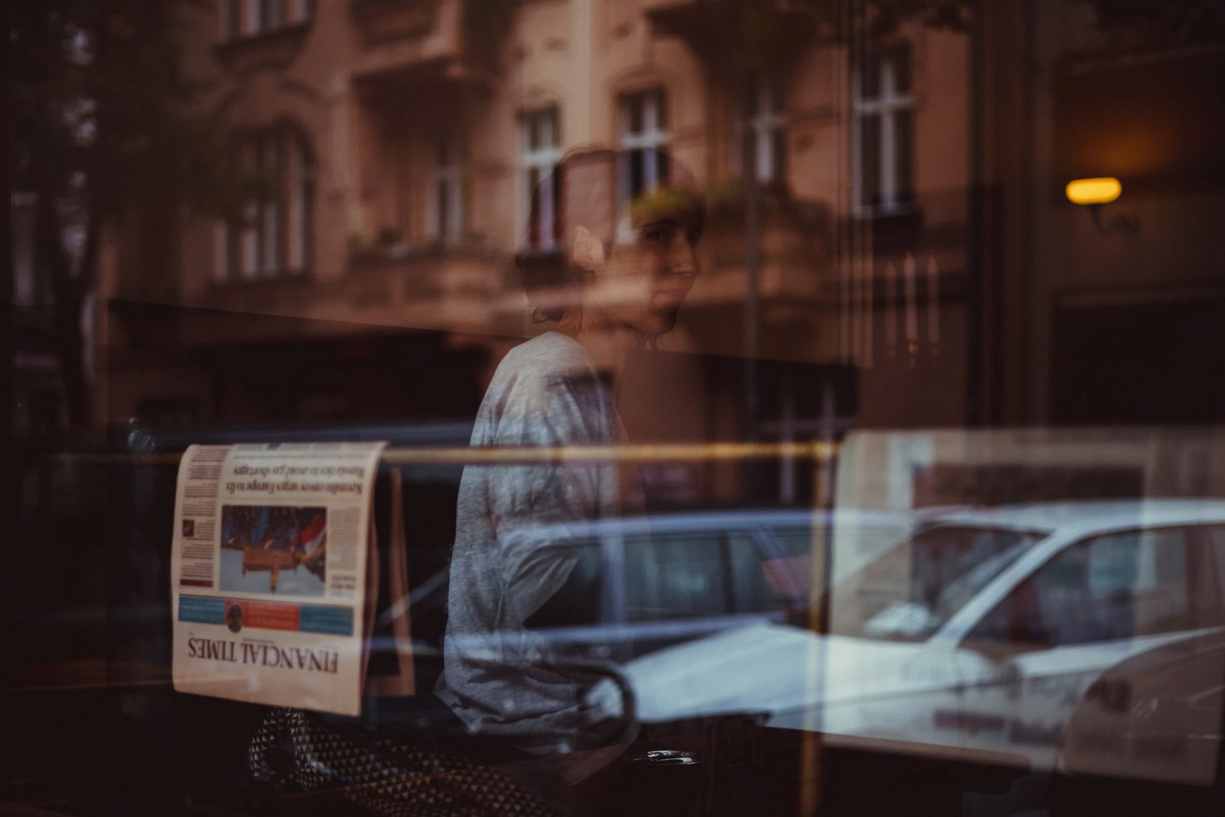 A reflection of a woman with short hair and a patterned shirt viewed through a window, with buildings and a newspaper visible behind the glass.