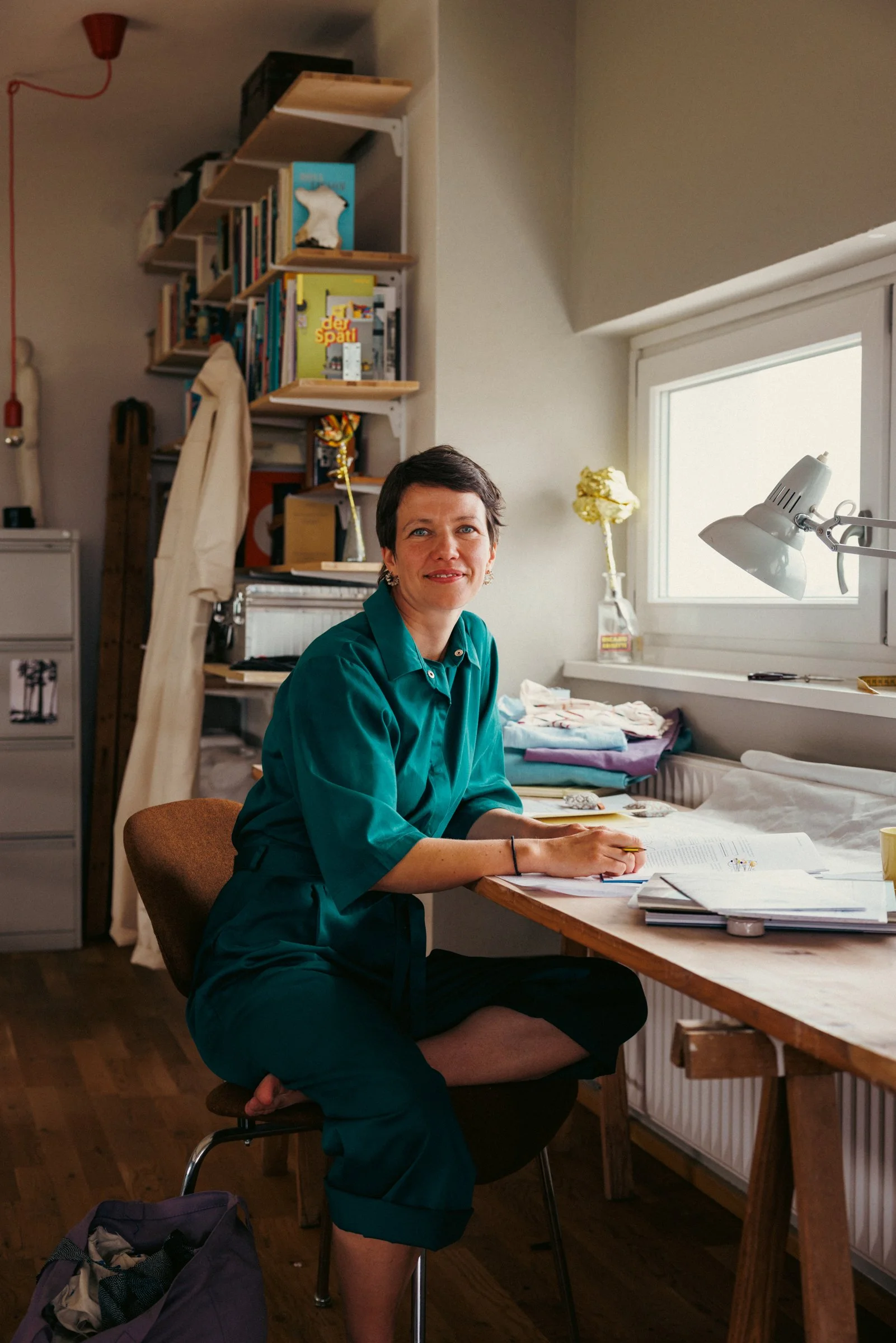 A woman with short dark hair, wearing a teal green shirt and dark pants, sits at a wooden table in a room with books, a window, and a lamp. She is smiling and looking at the camera.