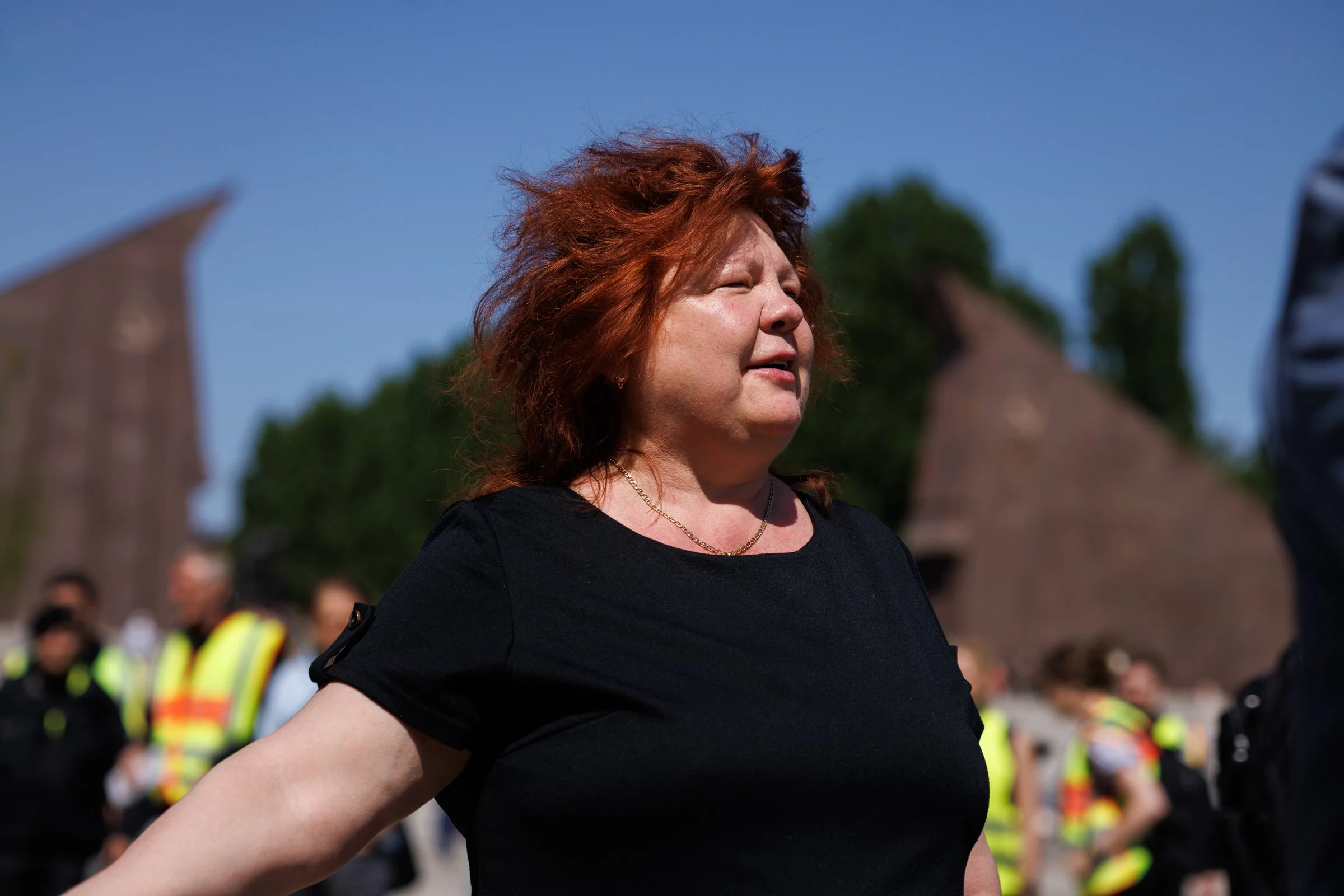 A woman with red hair and a black shirt stands outdoors during daytime, with people and police officers in reflective vests in the background.