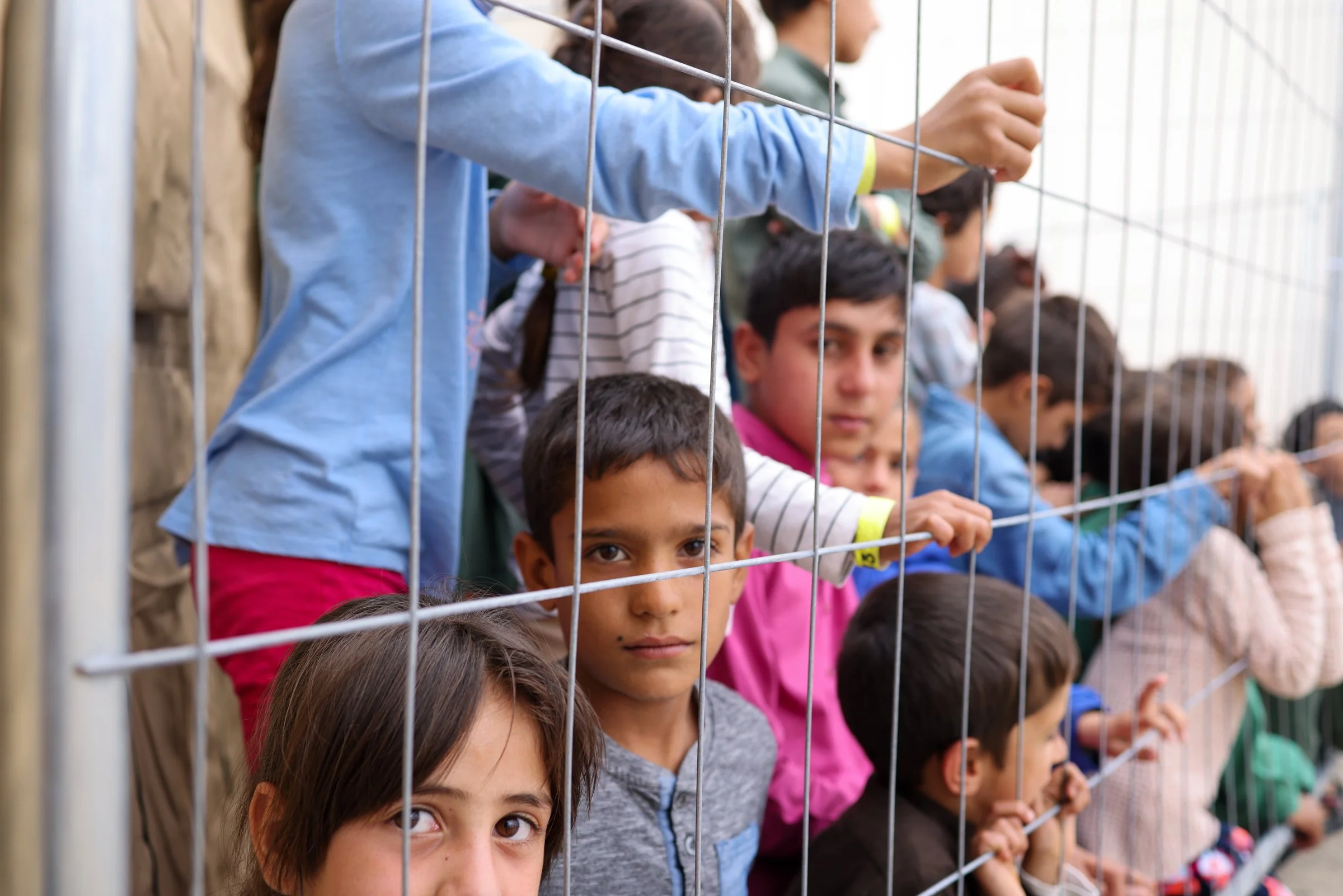 A group of Afghan children standing behind a wire fence, looking through it with curious expressions in Ramstein American Airbase in Germany.