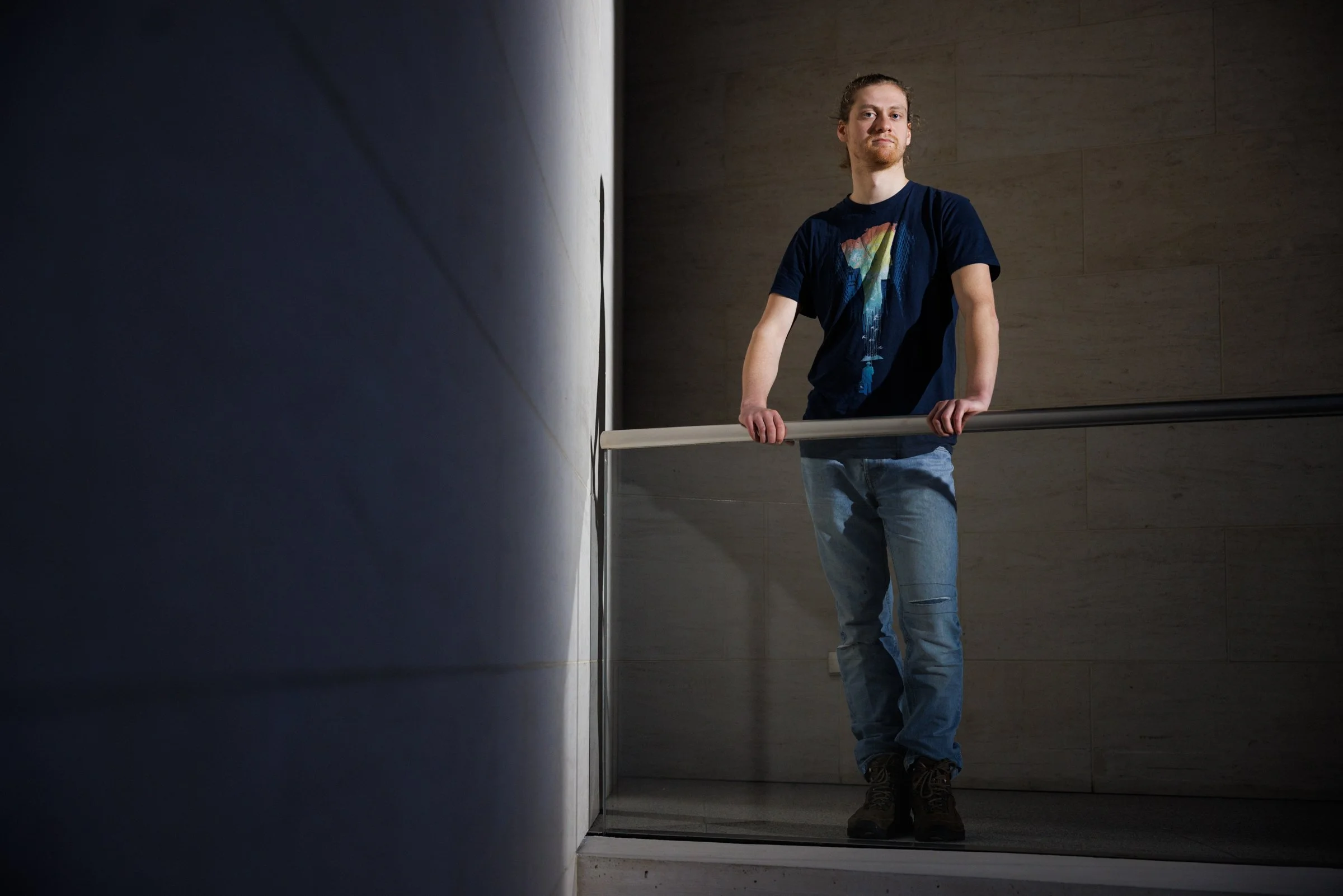 A man with long hair tied back, wearing a dark t-shirt with a rainbow-colored design and jeans, standing behind a handrail inside a modern building with tan tiled walls.
