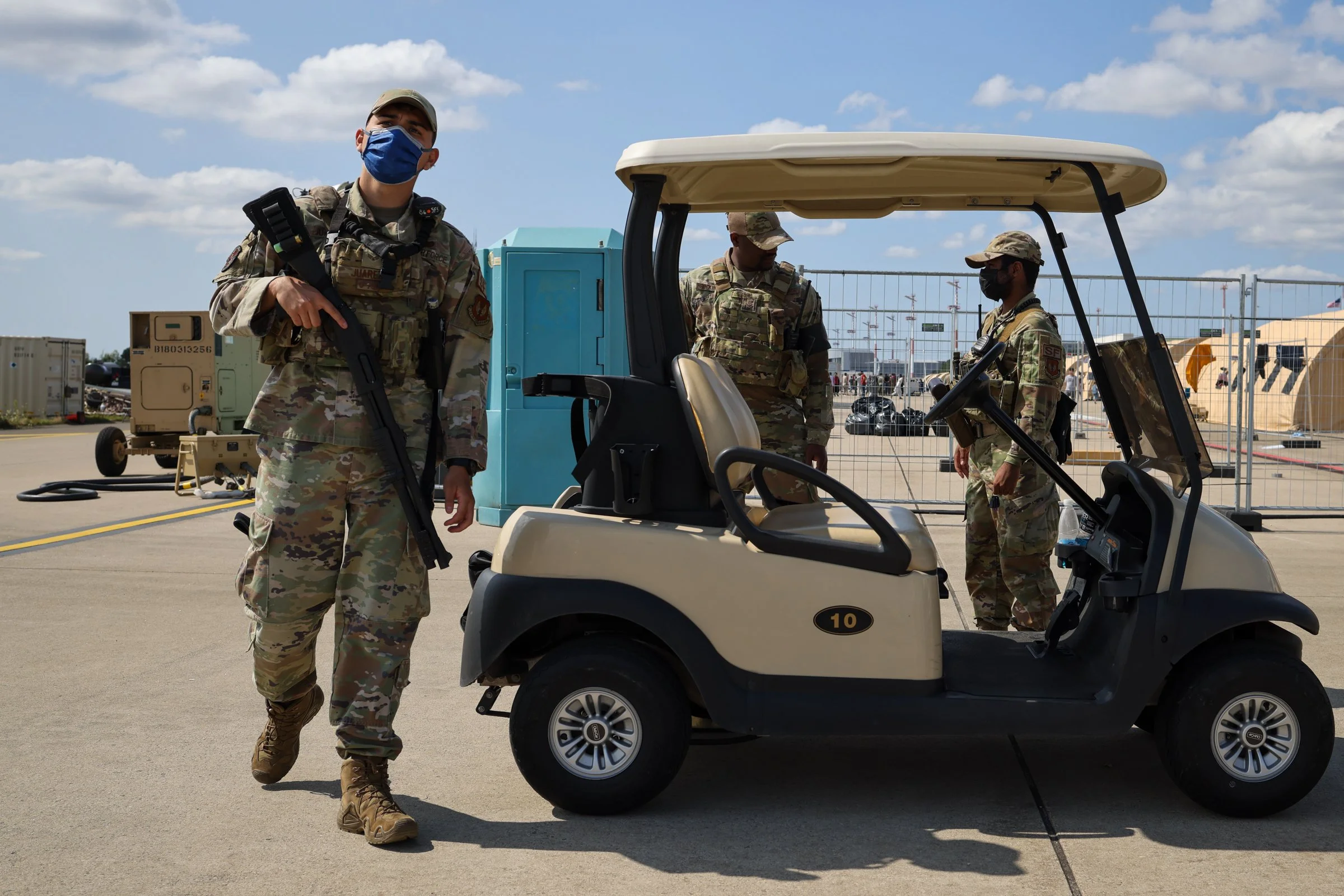 Three soldiers in camouflage uniforms and face masks standing next to a golf cart on an airport tarmac, with construction cranes and tents in the background in Ramstein American Airbase in Germany.