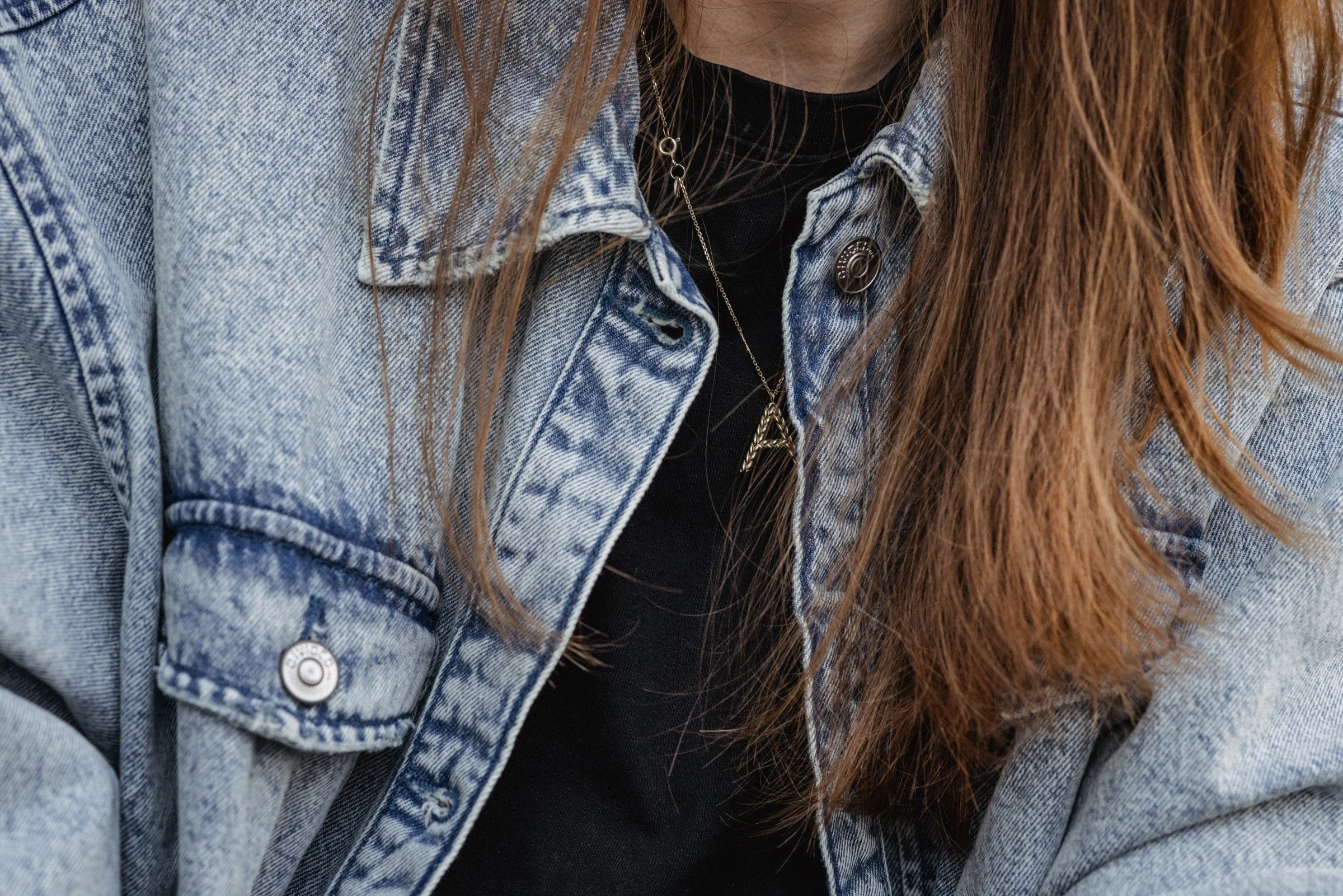 Close-up of a person wearing a light blue denim jacket over a black shirt, with long brown hair and layered necklaces.