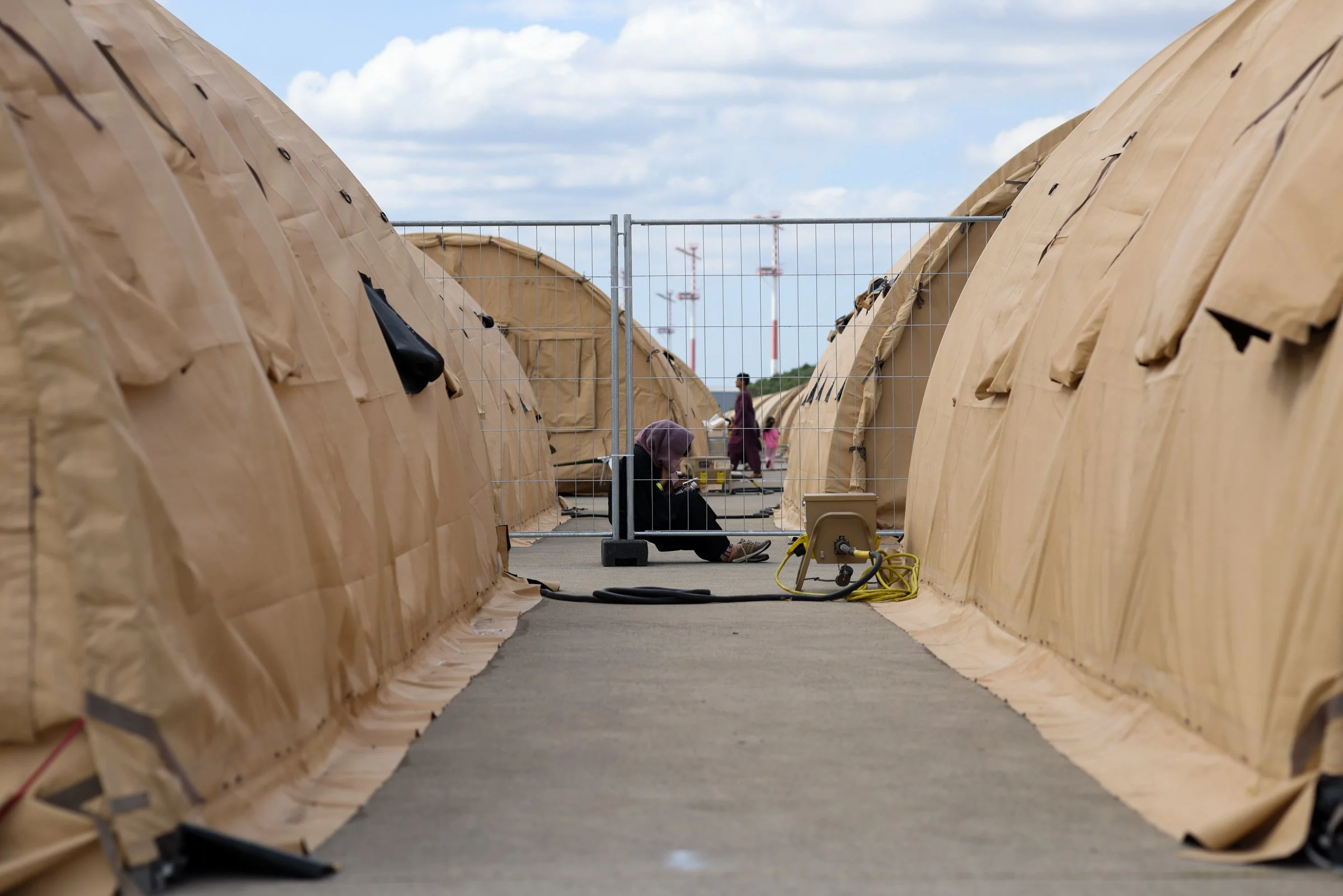 Outdoor scene showing large beige tents on either side, separated by a pathway, with a person sitting on the ground and another person walking in the background. The sky is partly cloudy in Ramstein American Airbase in Germany.