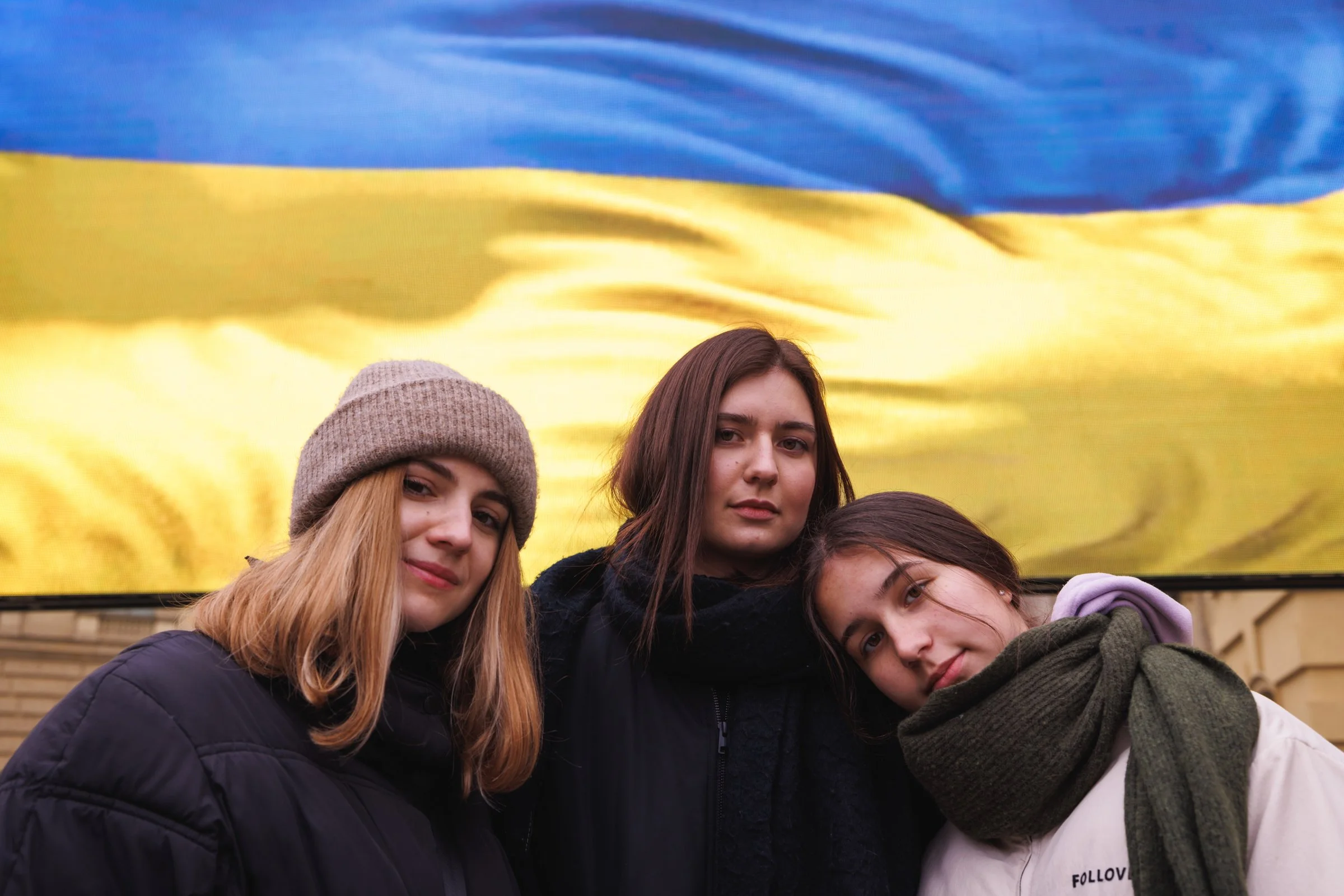 Group of young people standing together in front of a large Ukrainian flag, outdoors.