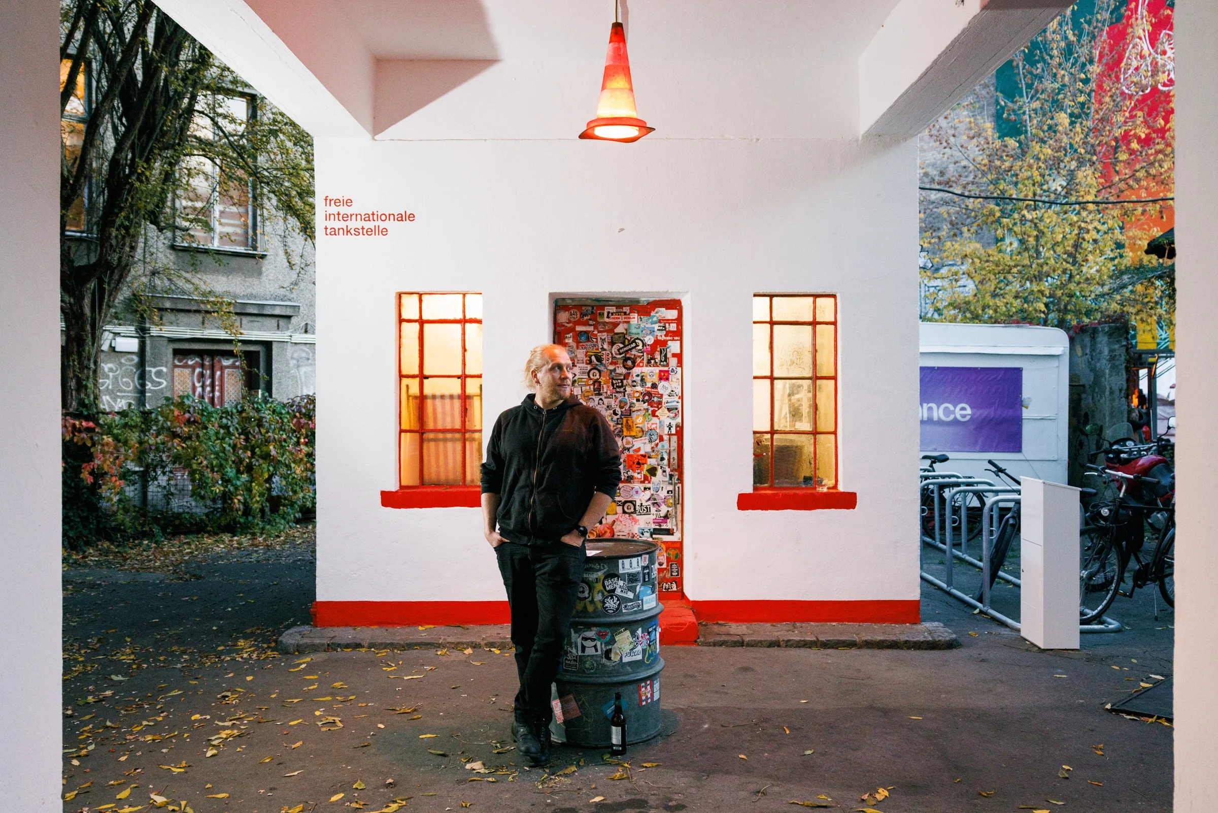 A man in black clothing with a beard and long hair tied back, standing outside under a small roof with a red-orange hanging light fixture. He is leaning on a painted barrel covered with stickers, next to two small orange windows on a white wall. Ther