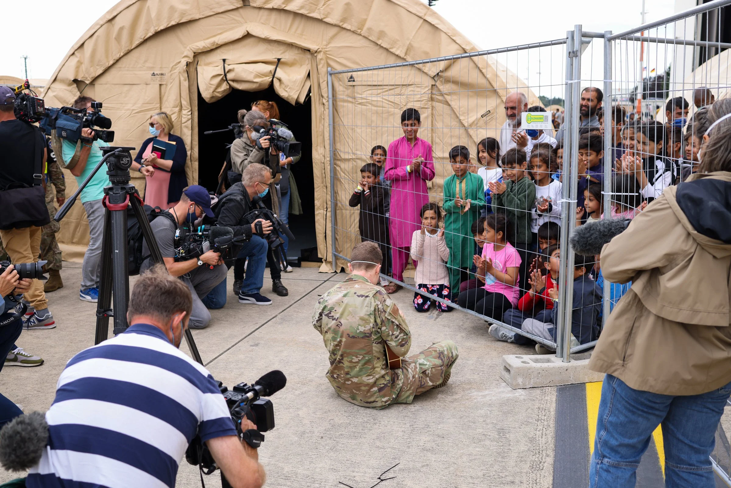 A group of Afghan children and adults behind a wire fence, some clapping and smiling, with a soldier kneeling on the ground in front of them during an outdoor event, many people filming or photographing  in Ramstein American Airbase in Germany.