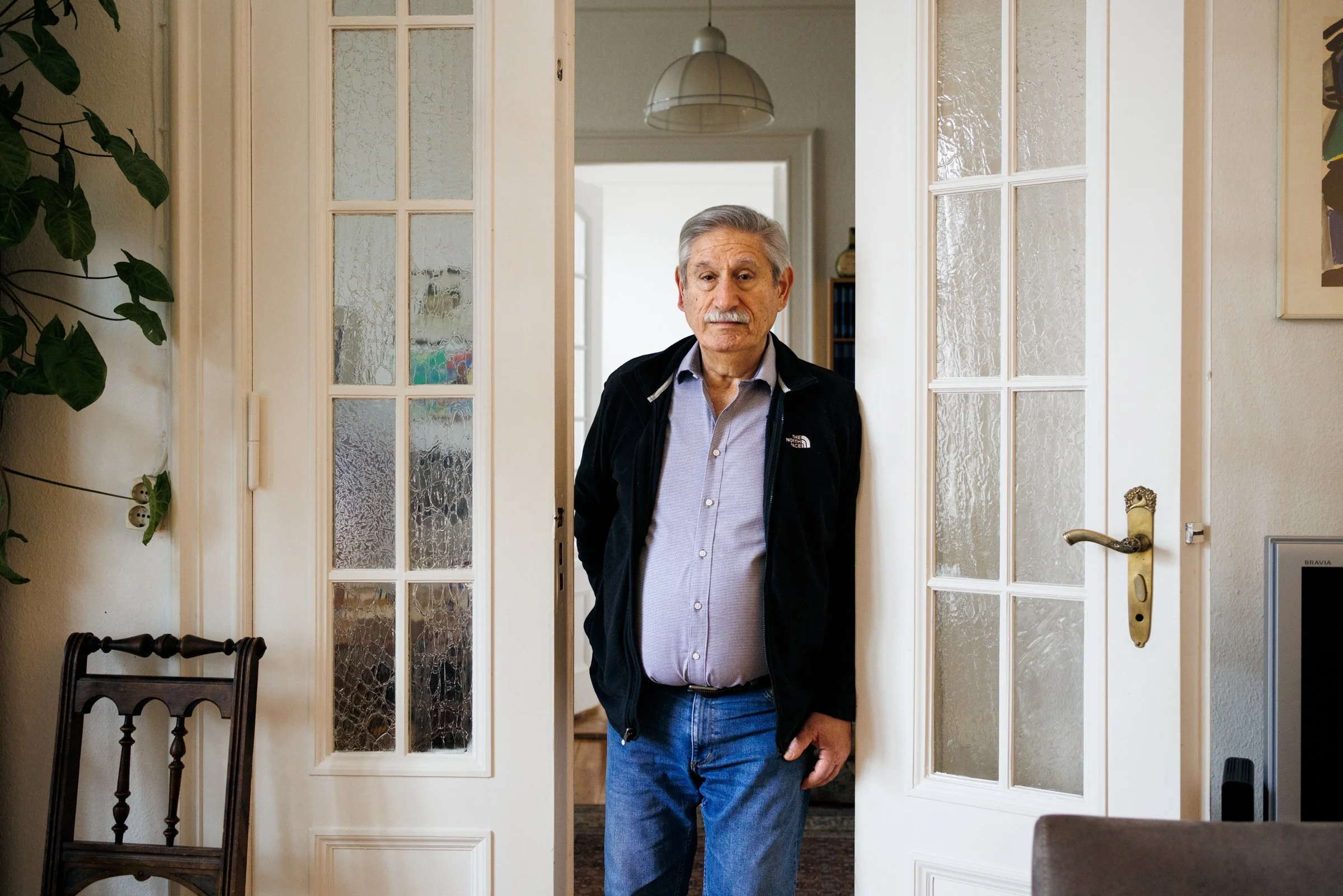 An elderly man with gray hair and a mustache standing in a doorway, wearing a light blue button-up shirt and a black jacket. The doorway is made of glass panes with textured glass and a gold-colored handle, leading to a room with white walls and a ce