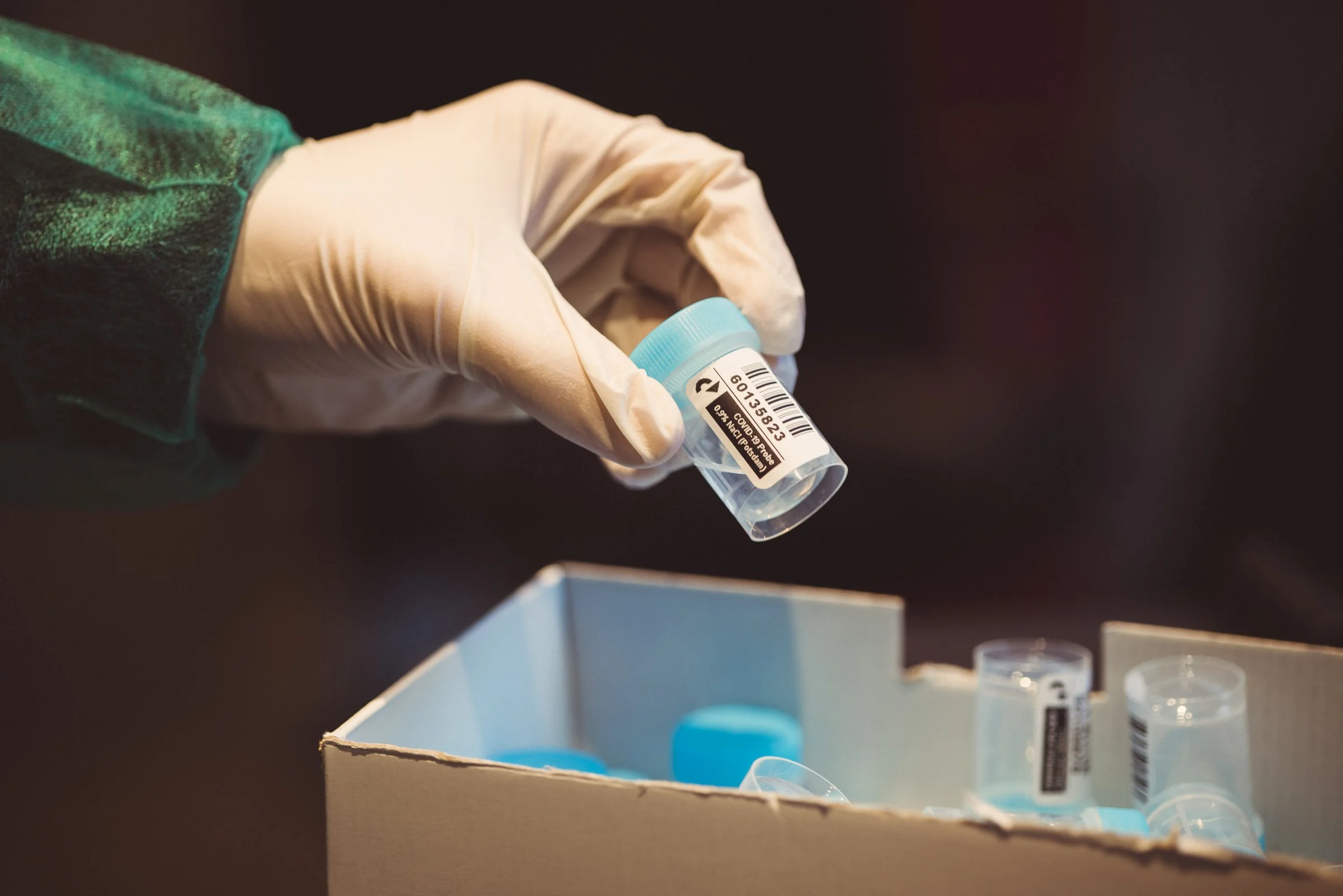 A gloved hand holding a small plastic container labeled with a barcode, with other similar containers and a cardboard box in the background.