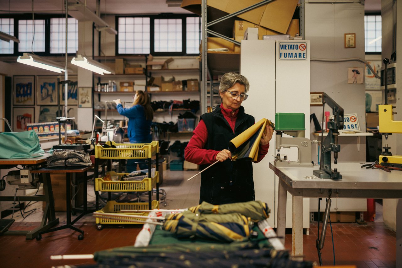 Woman artisan in a red vest inspecting fabric or components at a workstation in the Maglia workshop.