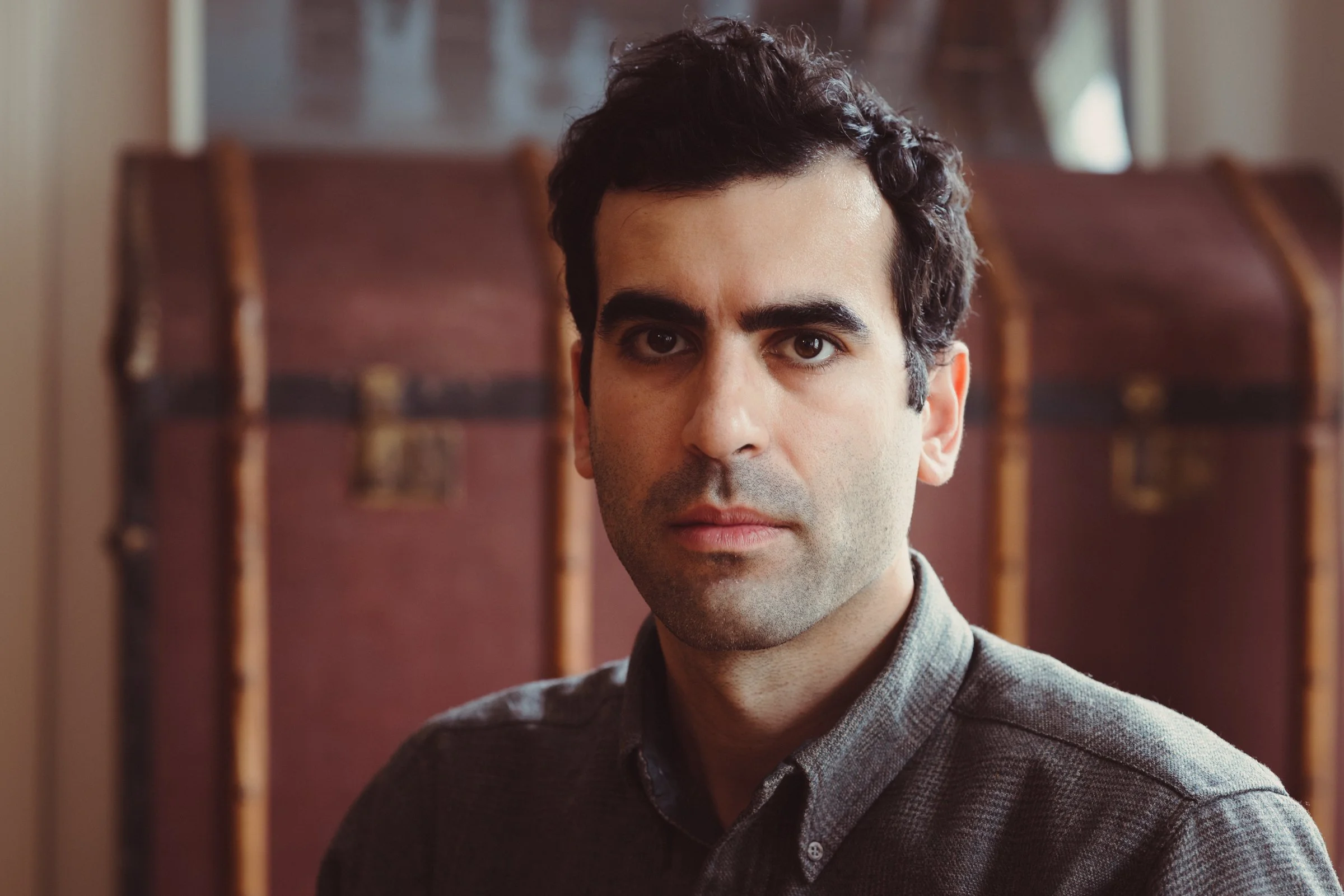 Aaron Benanav looking directly at the camera, partially obscured by a wooden object in the foreground, wearing a plaid shirt in an indoor setting.
