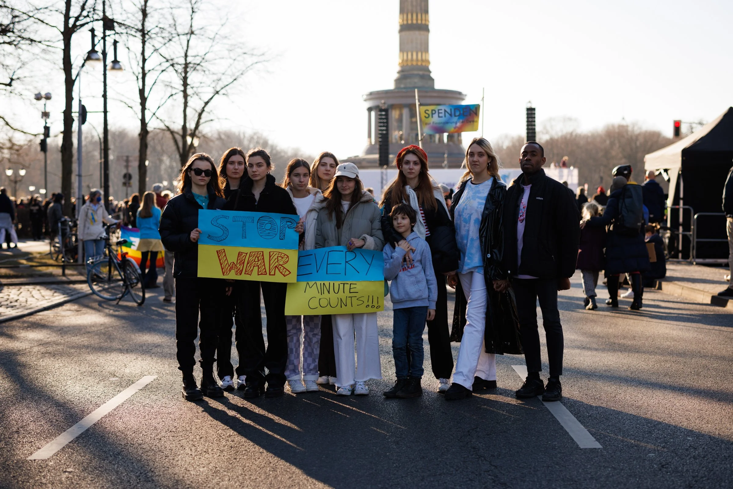 Group of young protesters holding signs protesting war during a demonstration in front of a landmark with a tall, decorated tower, possibly in a city park. The signs call for stopping war and highlight the importance of minute counts.