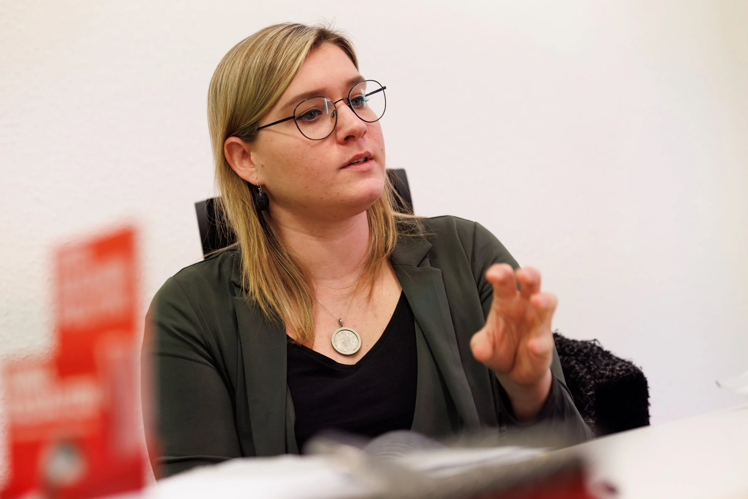 A woman with blonde hair and glasses sitting at a table, engaged in conversation, with a plain white wall in the background.