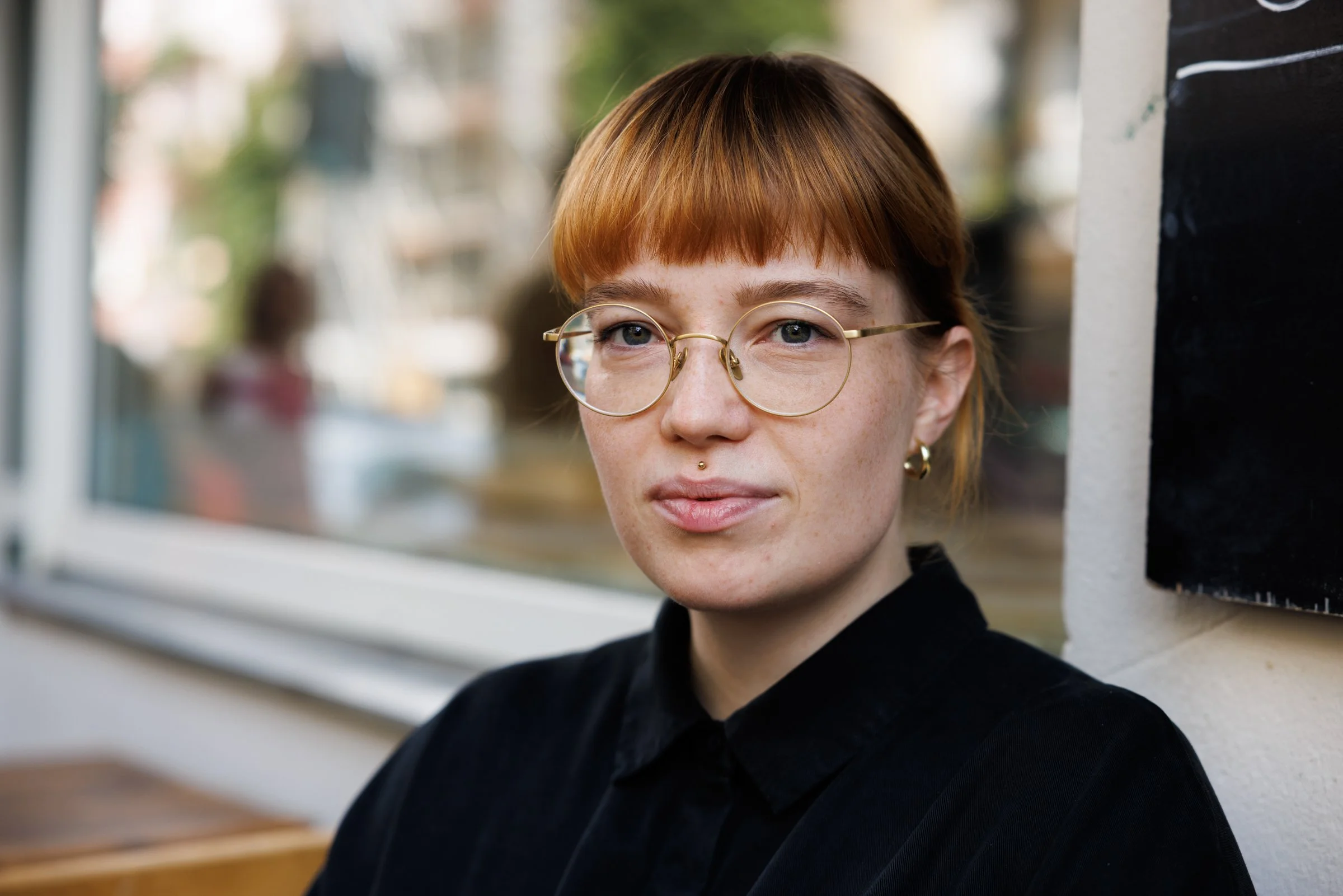 Close-up of a young woman with short red hair, wearing round glasses, black clothing, and a small facial piercing, looking out a window.