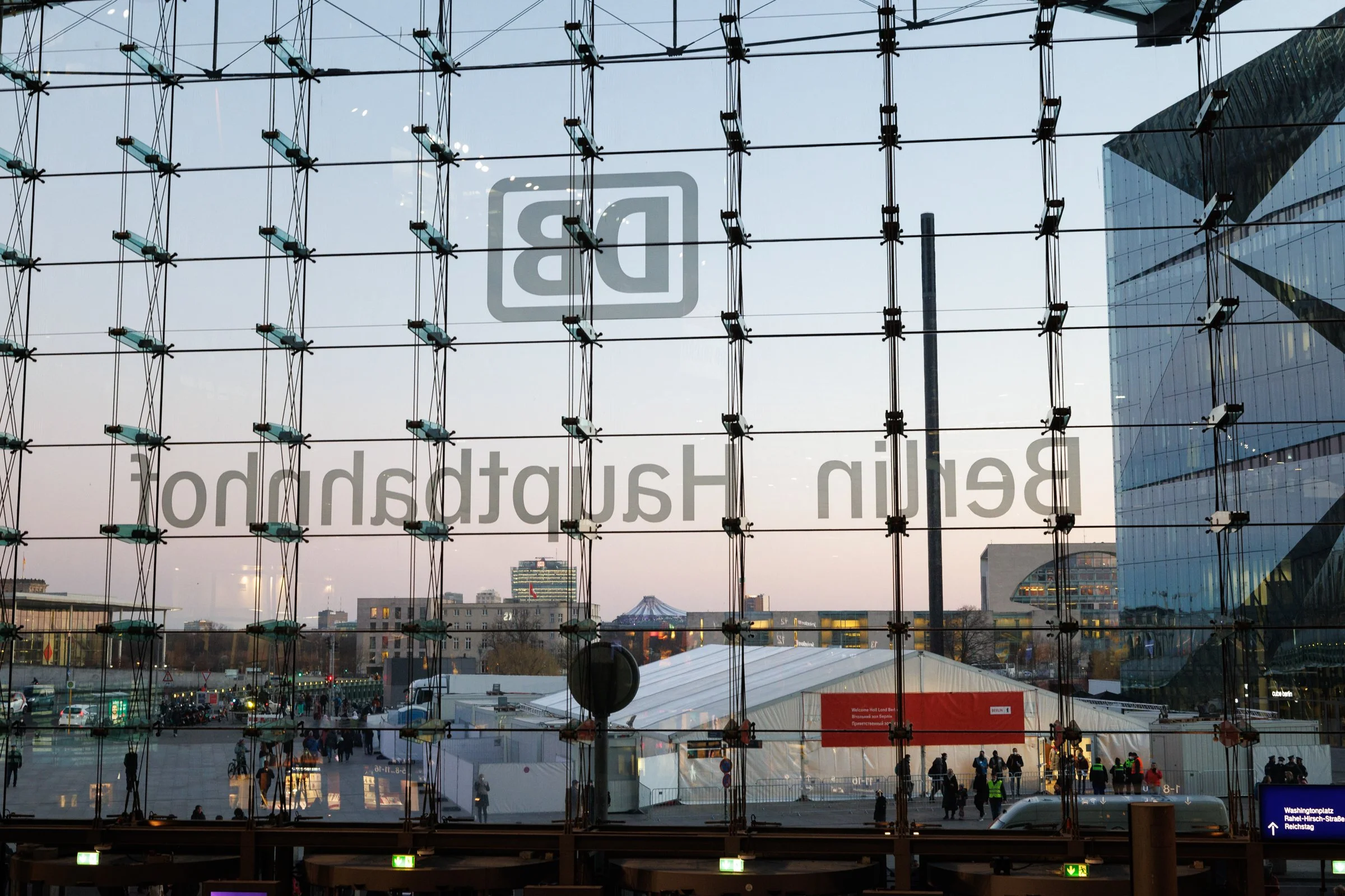 The image shows a large glass window with a reflection of an urban scene. Inside, there are many people walking, and outside, buildings and trees are visible. There is a sign in the lower right corner pointing to a street name, and some lighting fixt