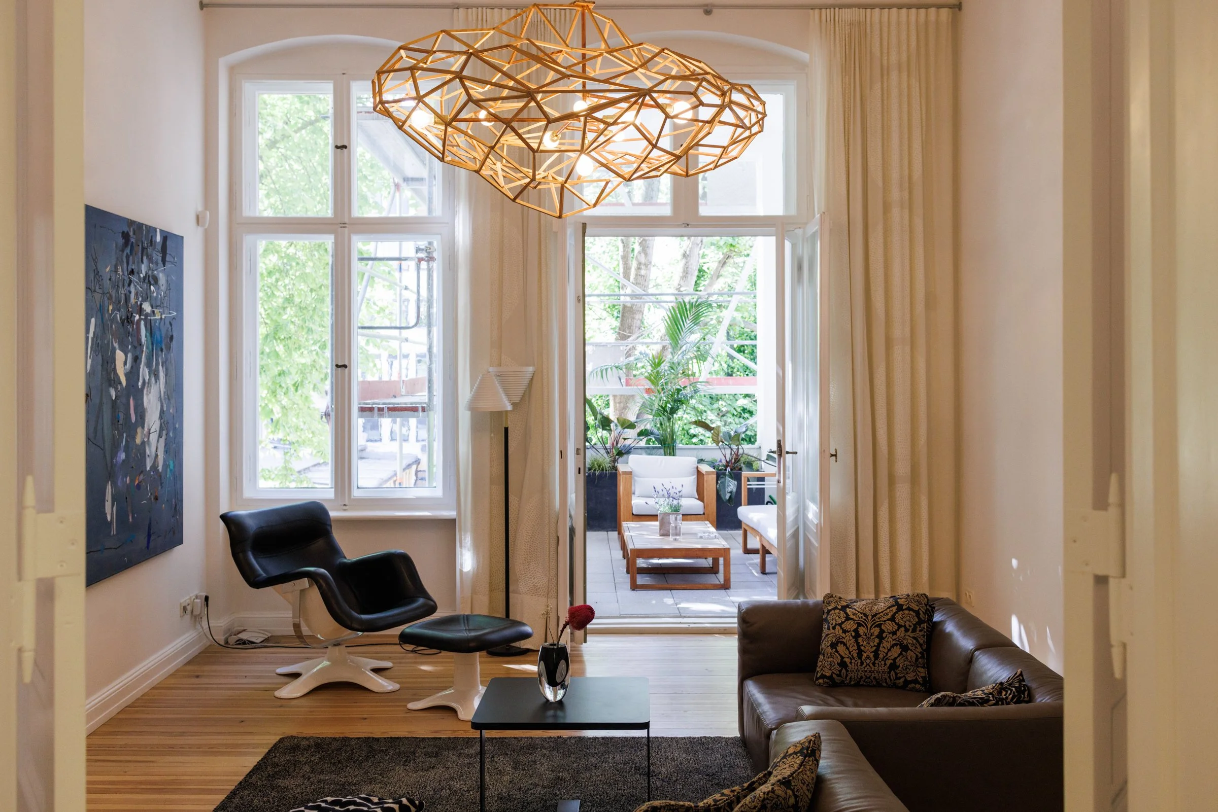Living room with a large geometric chandelier, black chair, leather sofa with patterned pillows, small black coffee table with a vase of flowers, and a balcony with outdoor furniture and greenery visible through large windows.