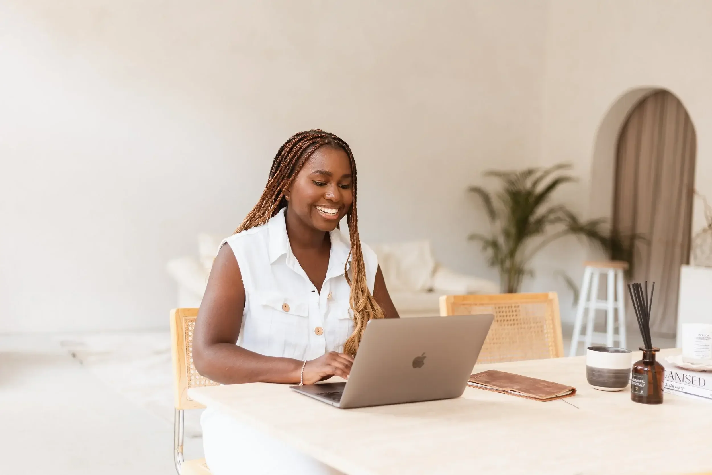 A woman with braided hair smiling while working on a silver laptop at a light-colored table in a bright, minimalist room with plants and decor.