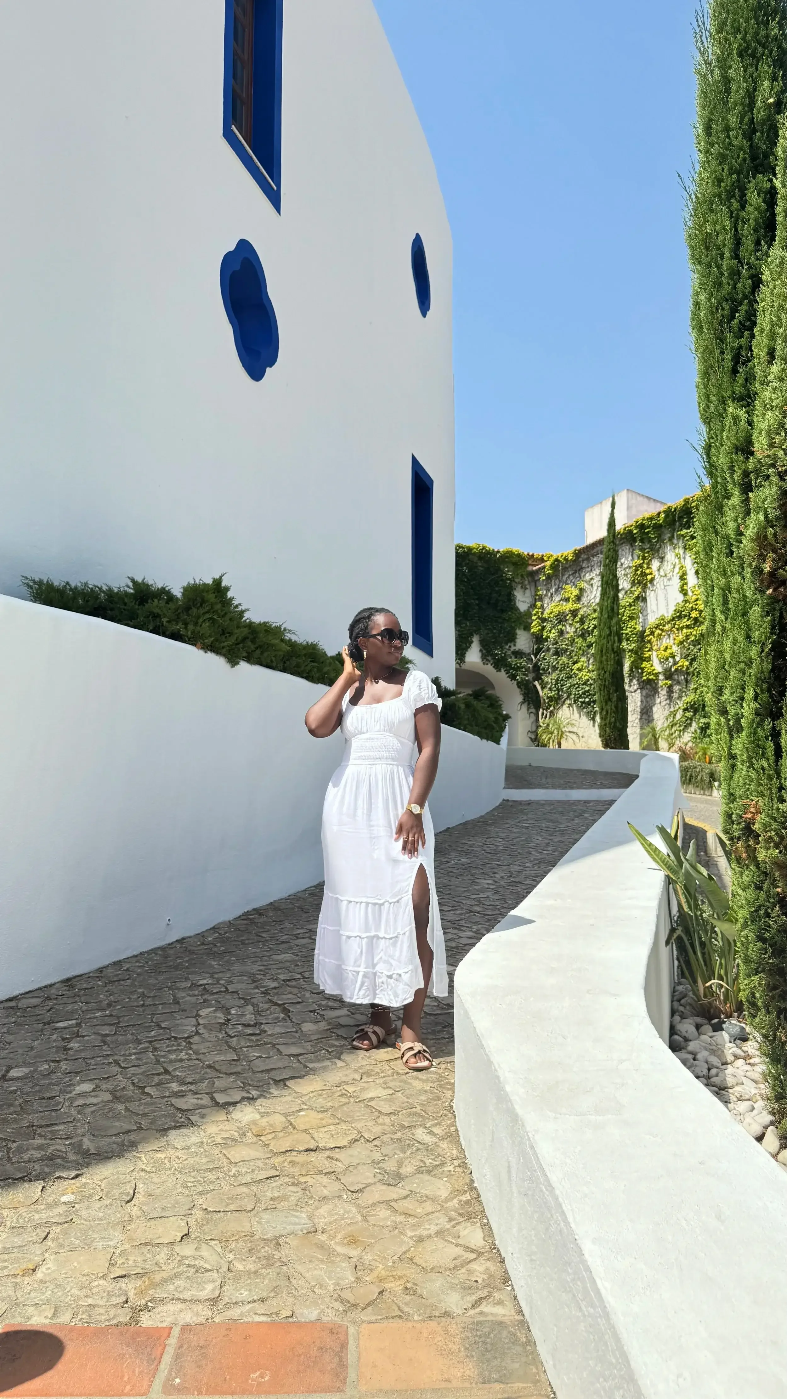 A woman in a white dress and sunglasses standing on a cobblestone path next to a white wall with blue windows and abstract blue wall art, surrounded by greenery and trees under a clear blue sky.