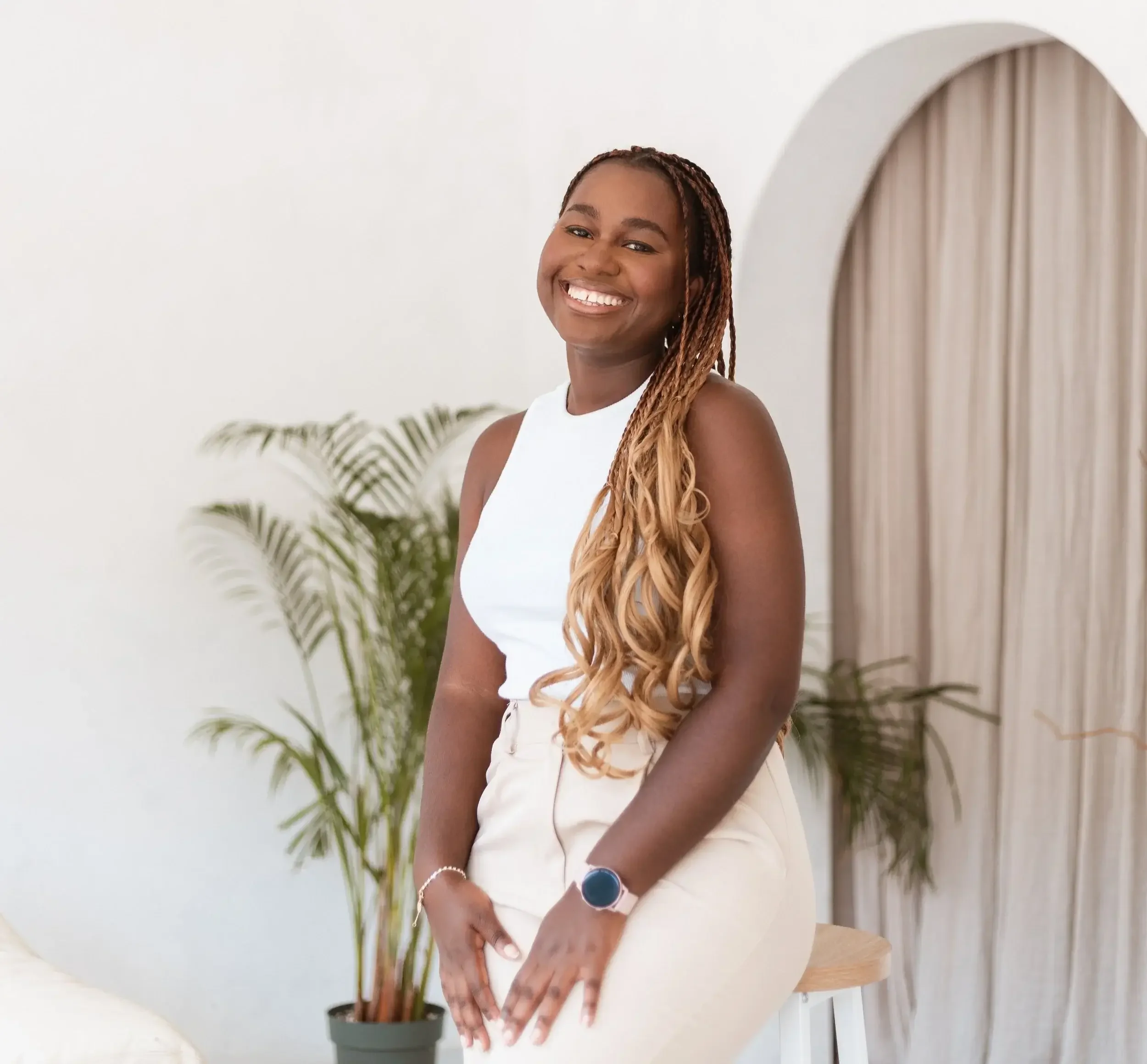 Smiling woman with braids wearing a white sleeveless top, standing indoors near a curved doorway and potted plants.