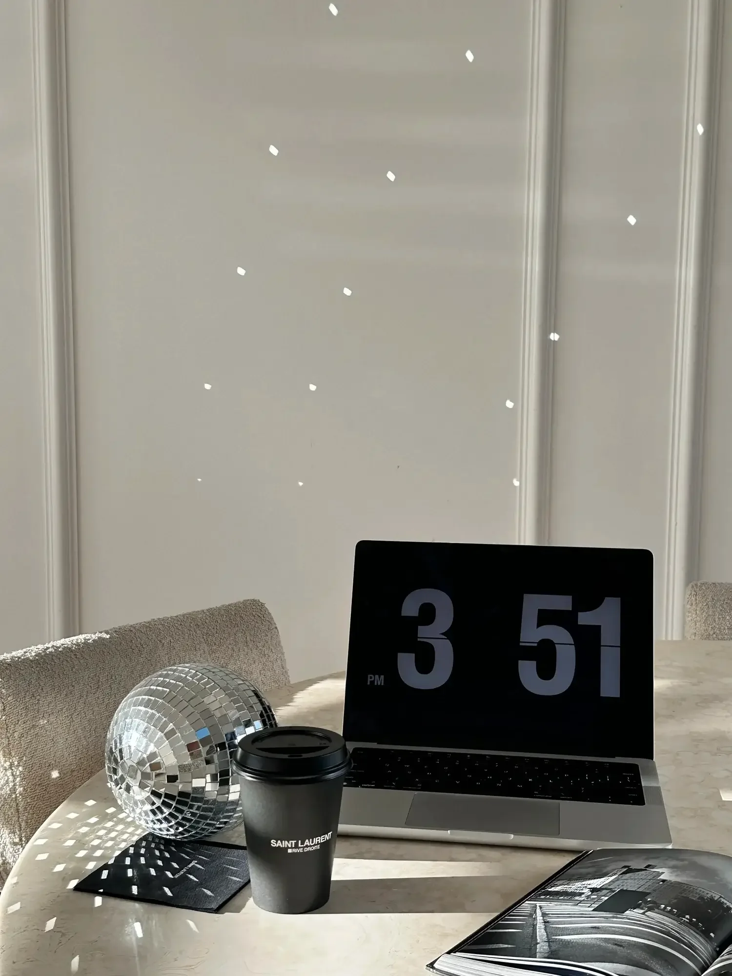 A modern desk setup with a silver laptop, a black Saint Laurent coffee cup, a disco ball, a black and white photo, and a large digital clock displaying 3:51 PM, with a white wall and ceiling with small recessed lights in the background.