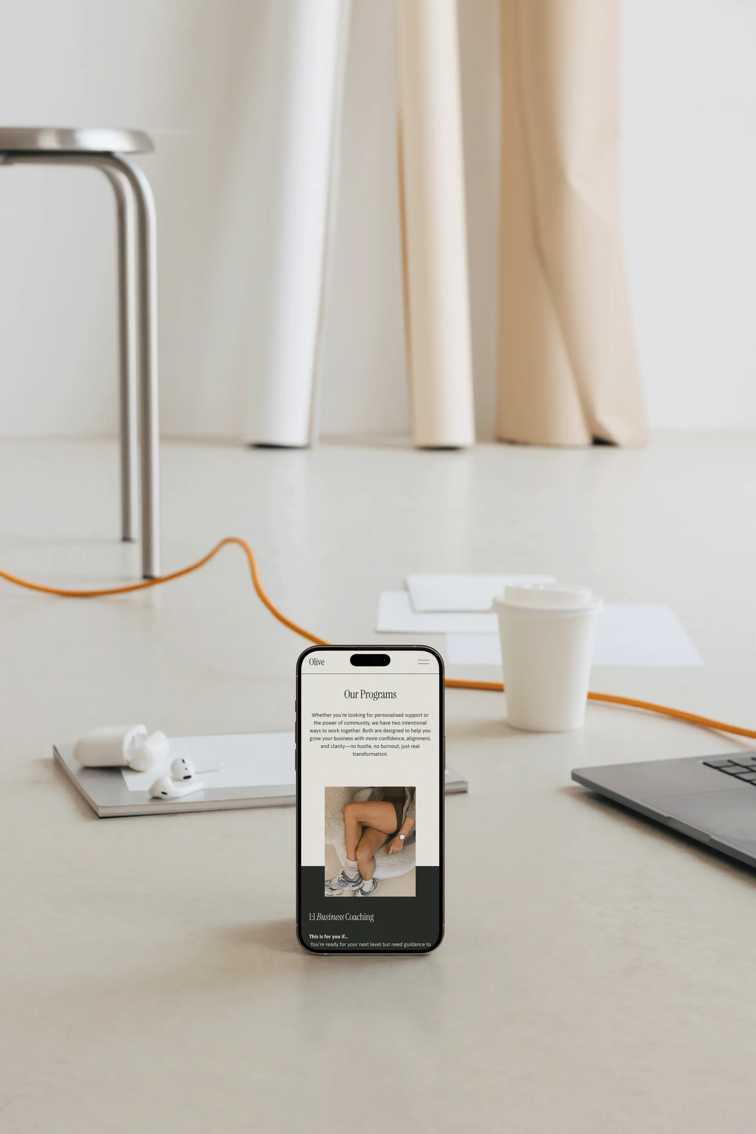 A white desk with wireless earbuds on a tray, a paper coffee cup, a smartphone displaying a website, a silver laptop, and a pair of white earbuds. In the background, beige curtains and a small table with a metal frame are visible.