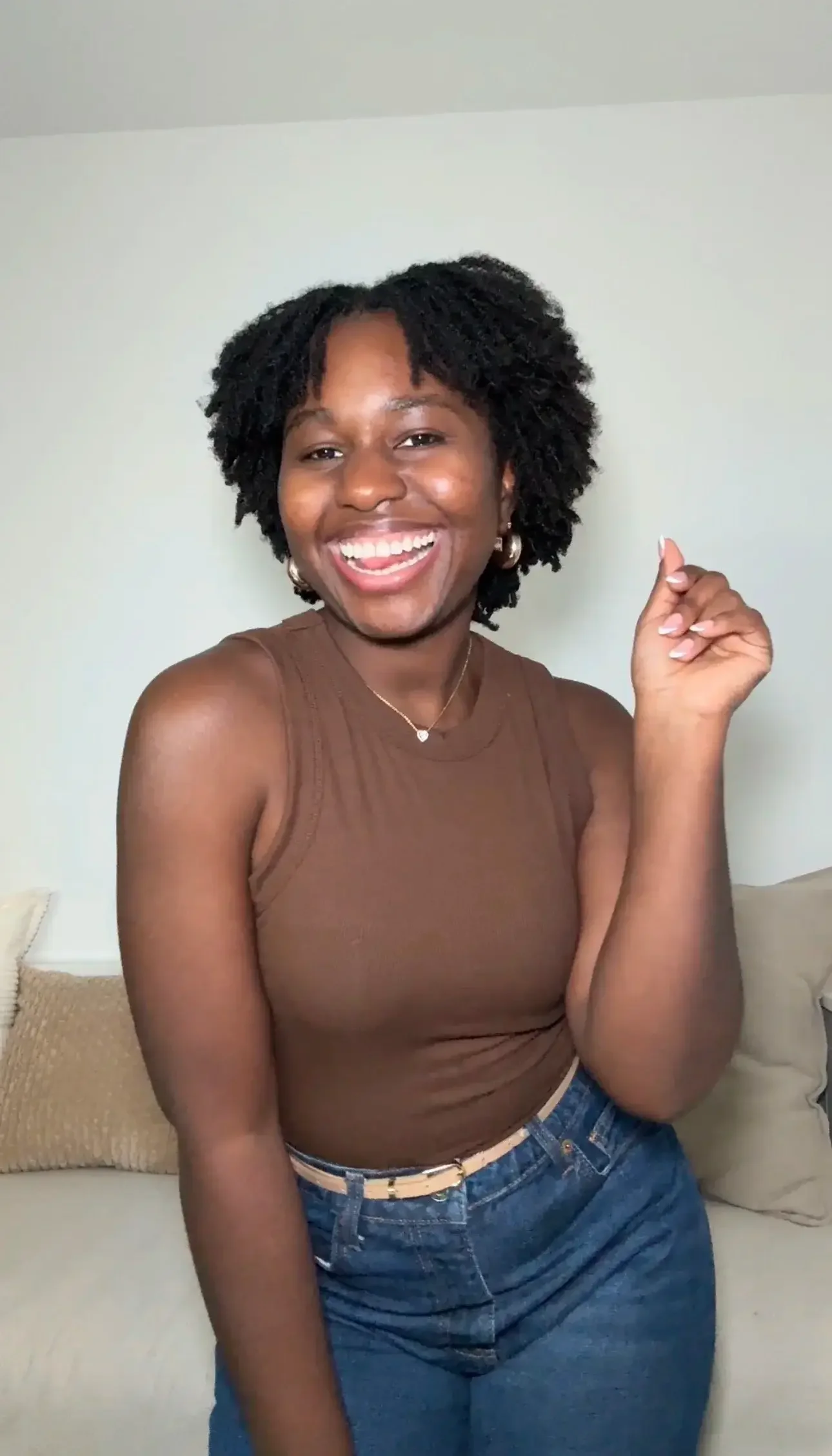 A woman with curly black hair wearing a brown tank top and blue jeans, smiling and showing her teeth, with her right hand raised near her face, sitting on a beige cushion in front of a white wall.