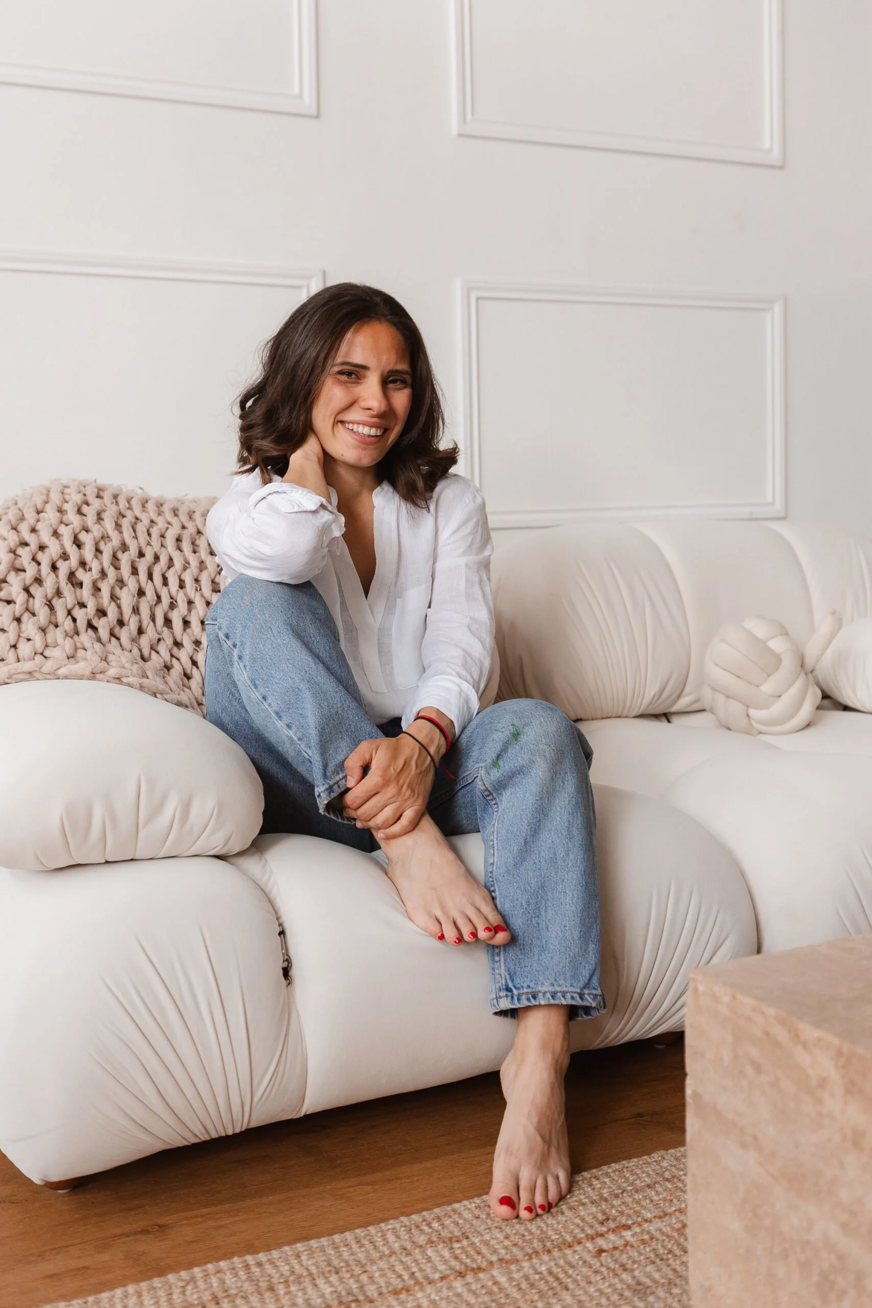 A woman with shoulder-length brown hair, wearing a white blouse and blue jeans, sitting on a white sofa, smiling warmly at the camera, with a cozy woven throw blanket behind her, in a bright, modern living room.