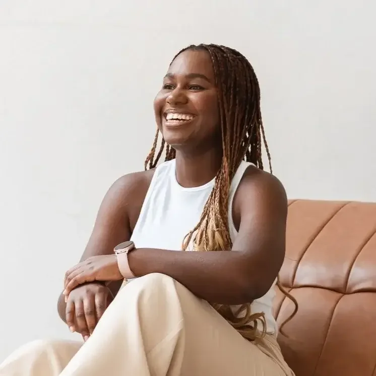 A woman with dreadlocks sitting on a tan couch, smiling and wearing a white sleeveless top and a watch.
