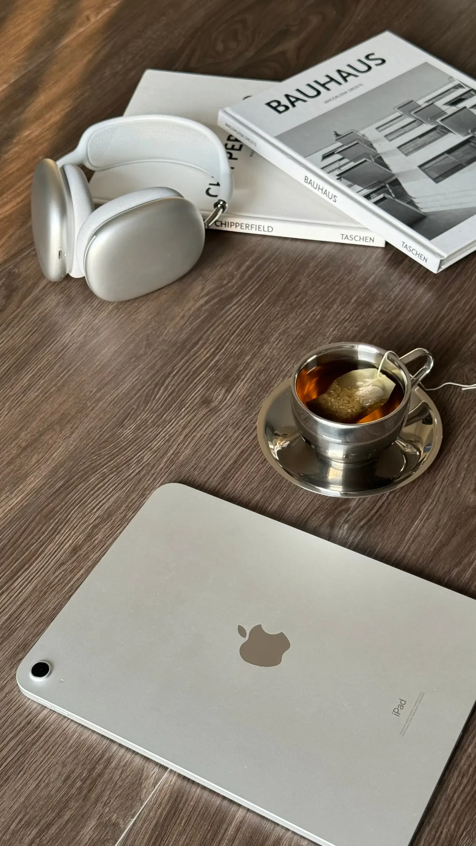 A silver iPad on a wooden table, a cup of tea with a tea bag inside, and two magazines or books about Bauhaus architecture.