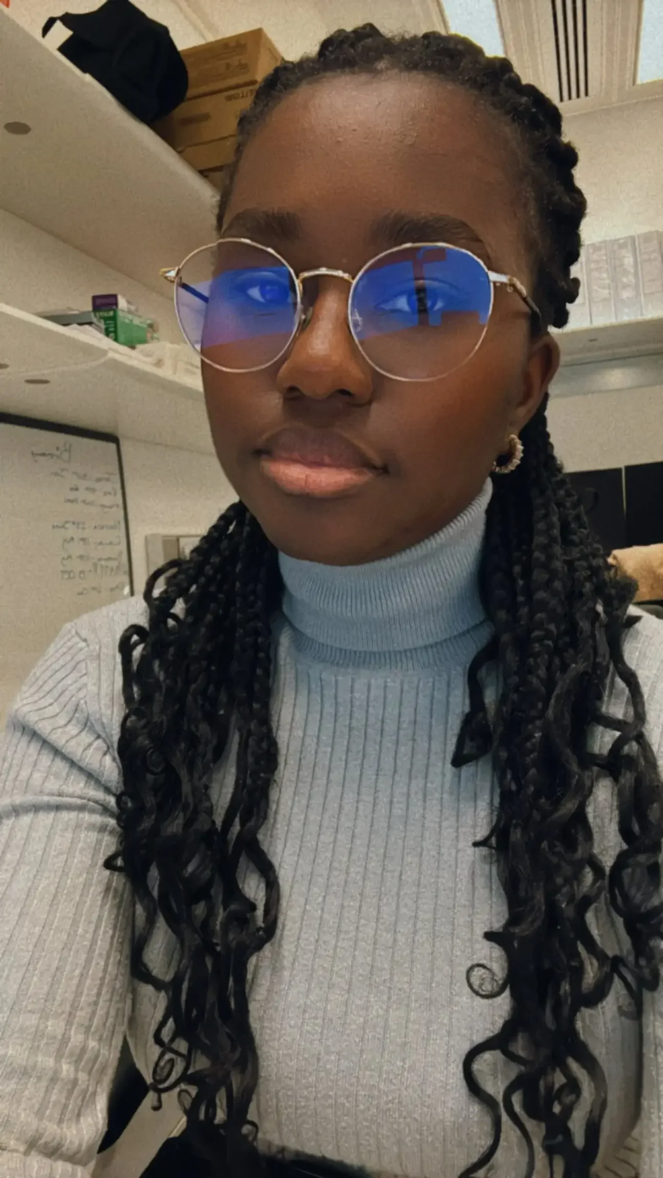 A woman with dark braided hair wearing round glasses with blue reflective lenses and a gray turtleneck sweater, sitting indoors with shelves and boxes in the background.