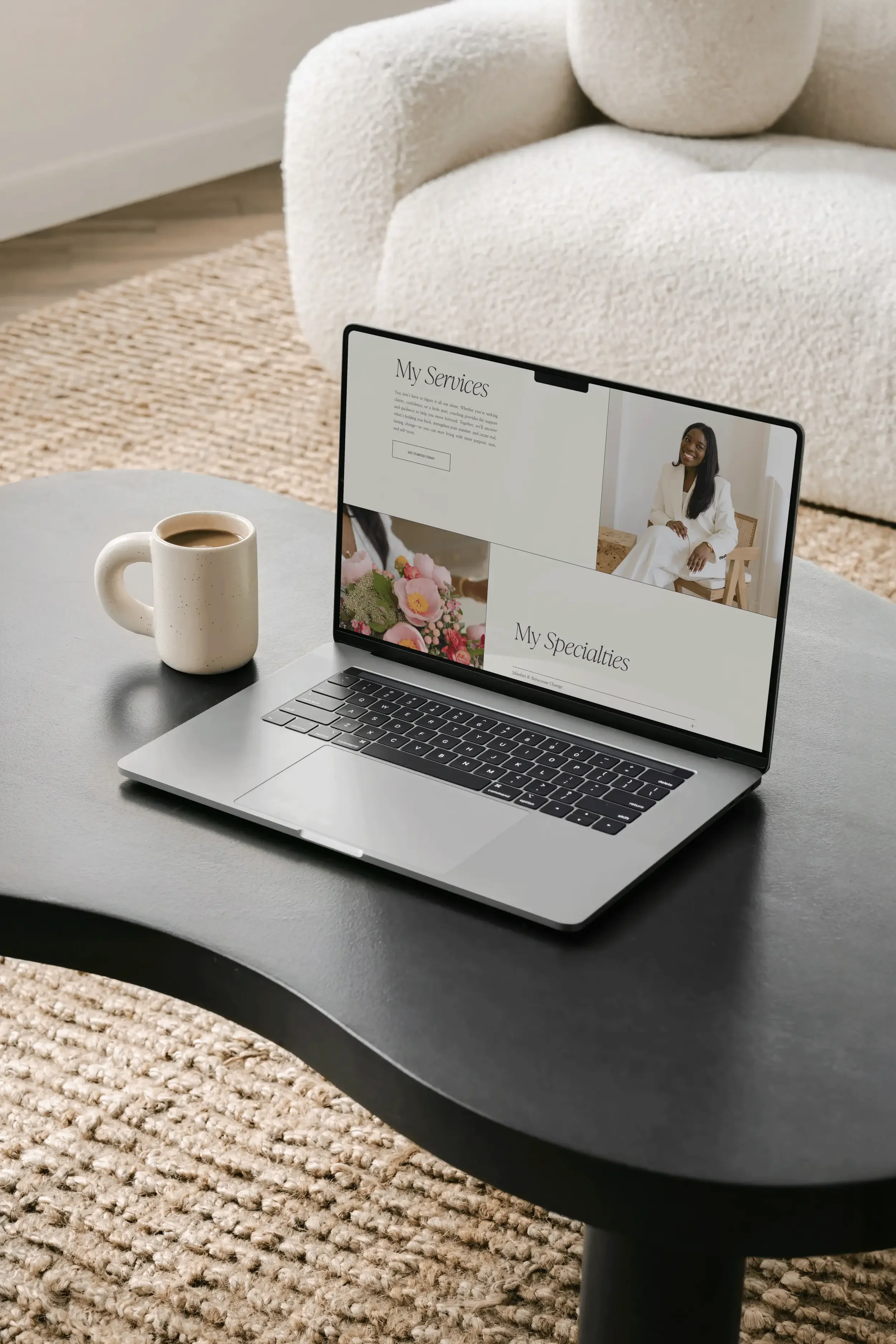 A gray table holding a silver laptop and a white mug filled with coffee, with a beige textured carpet and a white sofa in the background.