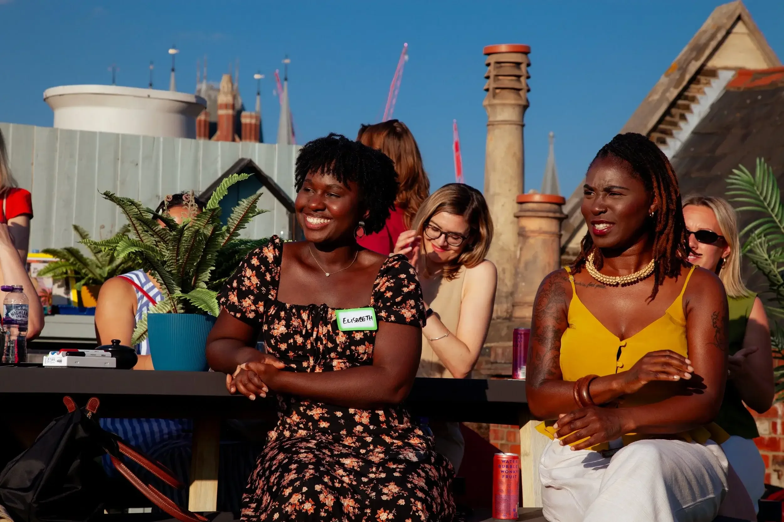 A group of diverse smiling women sitting at a table outdoors during daytime, with a cityscape and blue sky in the background.