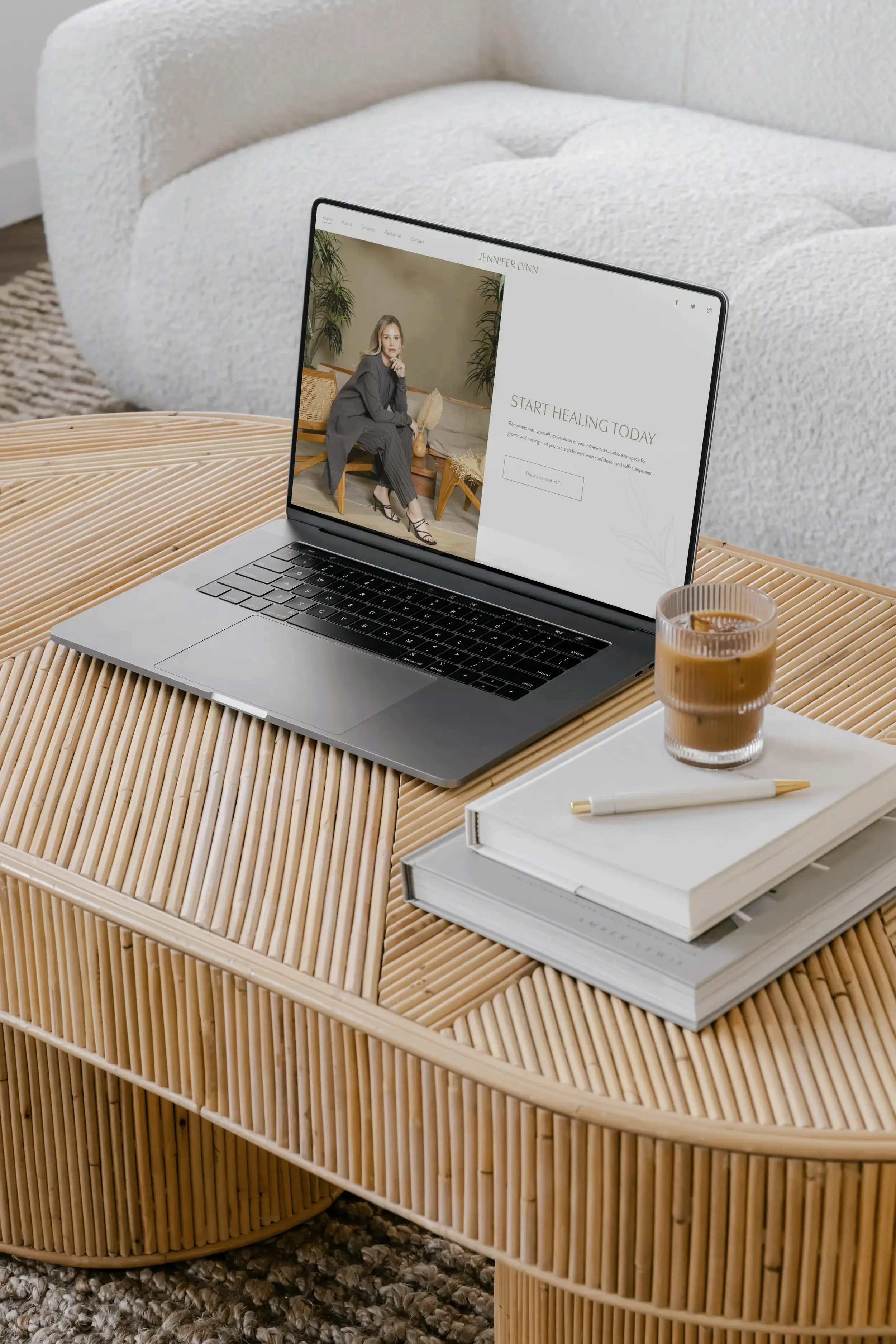 A laptop on a wooden table showing a website with a woman sitting and advertising healing services. Next to the laptop is a glass of iced coffee, a pen, and closed notebooks. The background features a white textured sofa and a gray carpet.