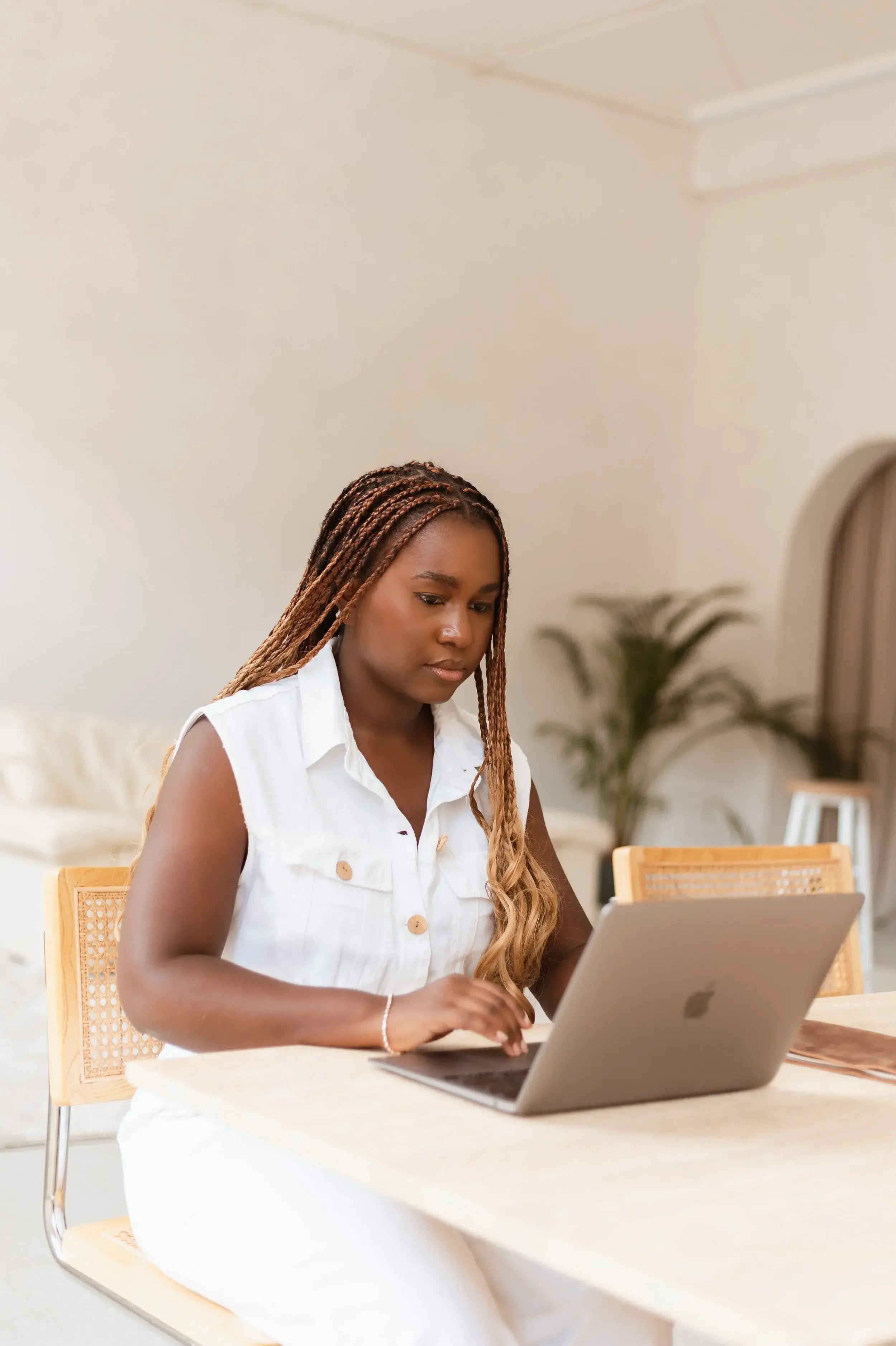 A woman with braided hair wearing a white sleeveless shirt working on a laptop at a table in a bright room with beige walls and a potted plant in the background.