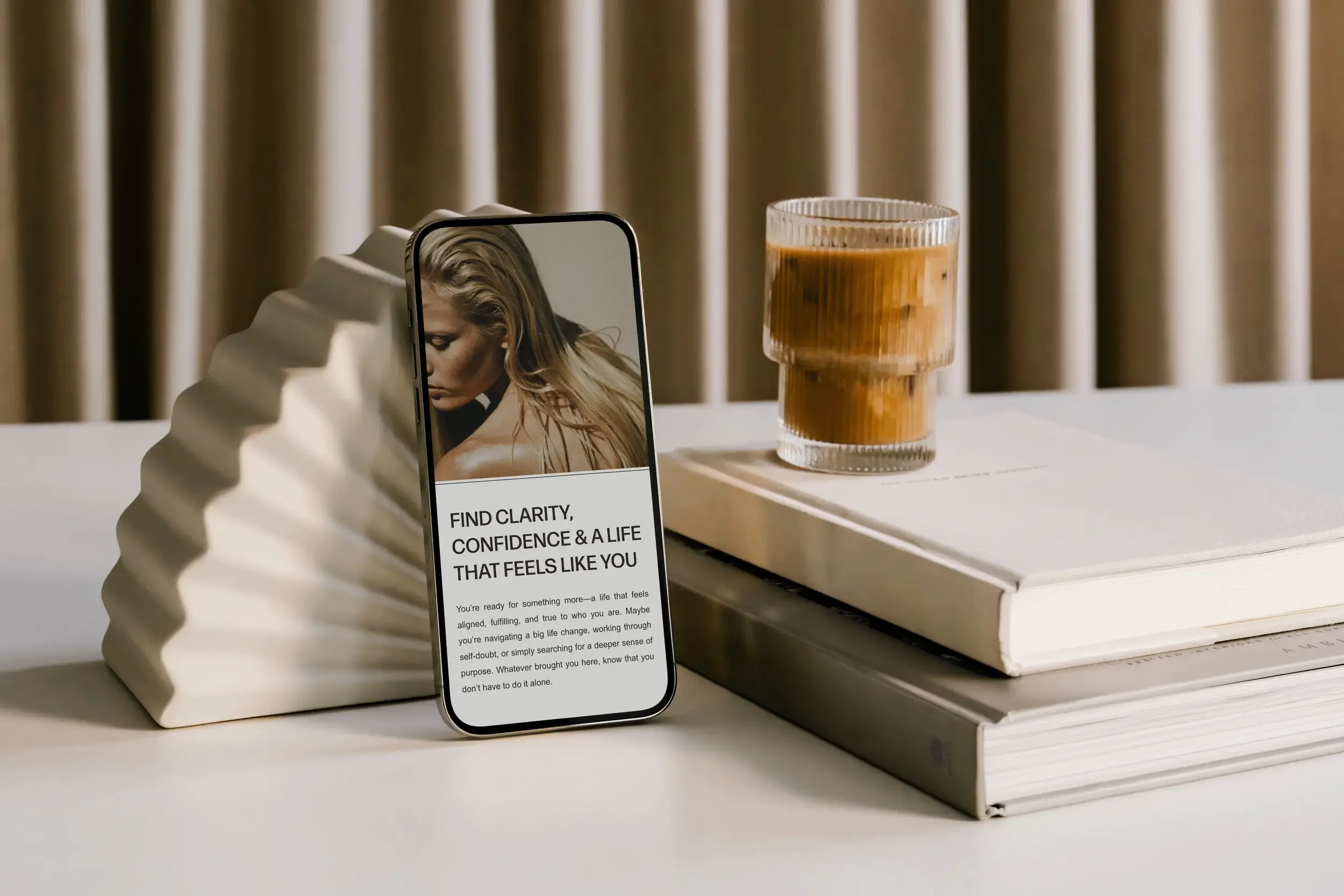A smartphone leaning on a white, wavy-shaped ceramic object on a white shelf. The phone screen displays text about finding clarity, confidence, and a life that feels aligned. Behind the display, there is a glass of iced coffee and a closed book.