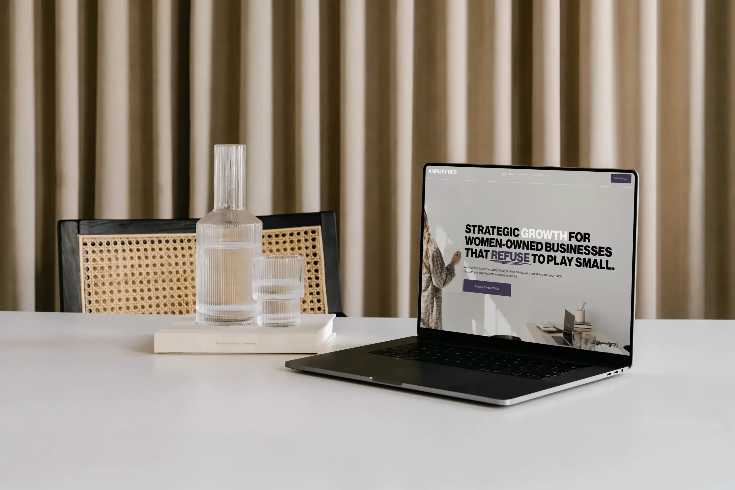 A workspace with a laptop displaying a website about strategic growth for women-owned businesses, a white table with decorative vases, and a woven chair in front of a curtain.